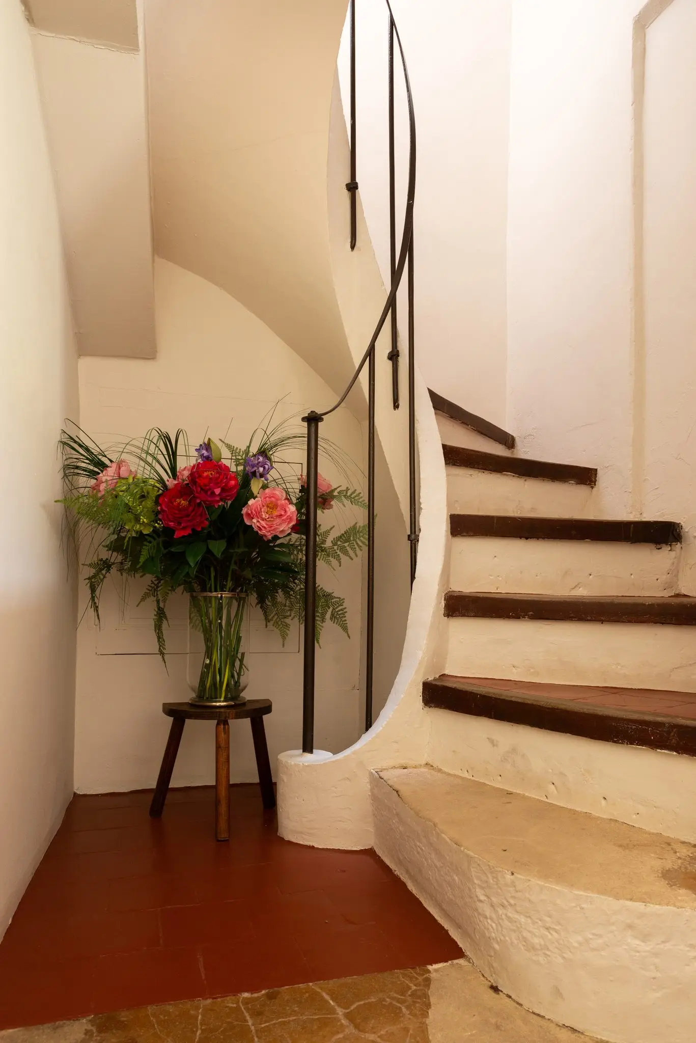 Escalier tournant en pierre blanchie à rampe de fer, bouquet de pivoines sur un tabouret au pied des marches