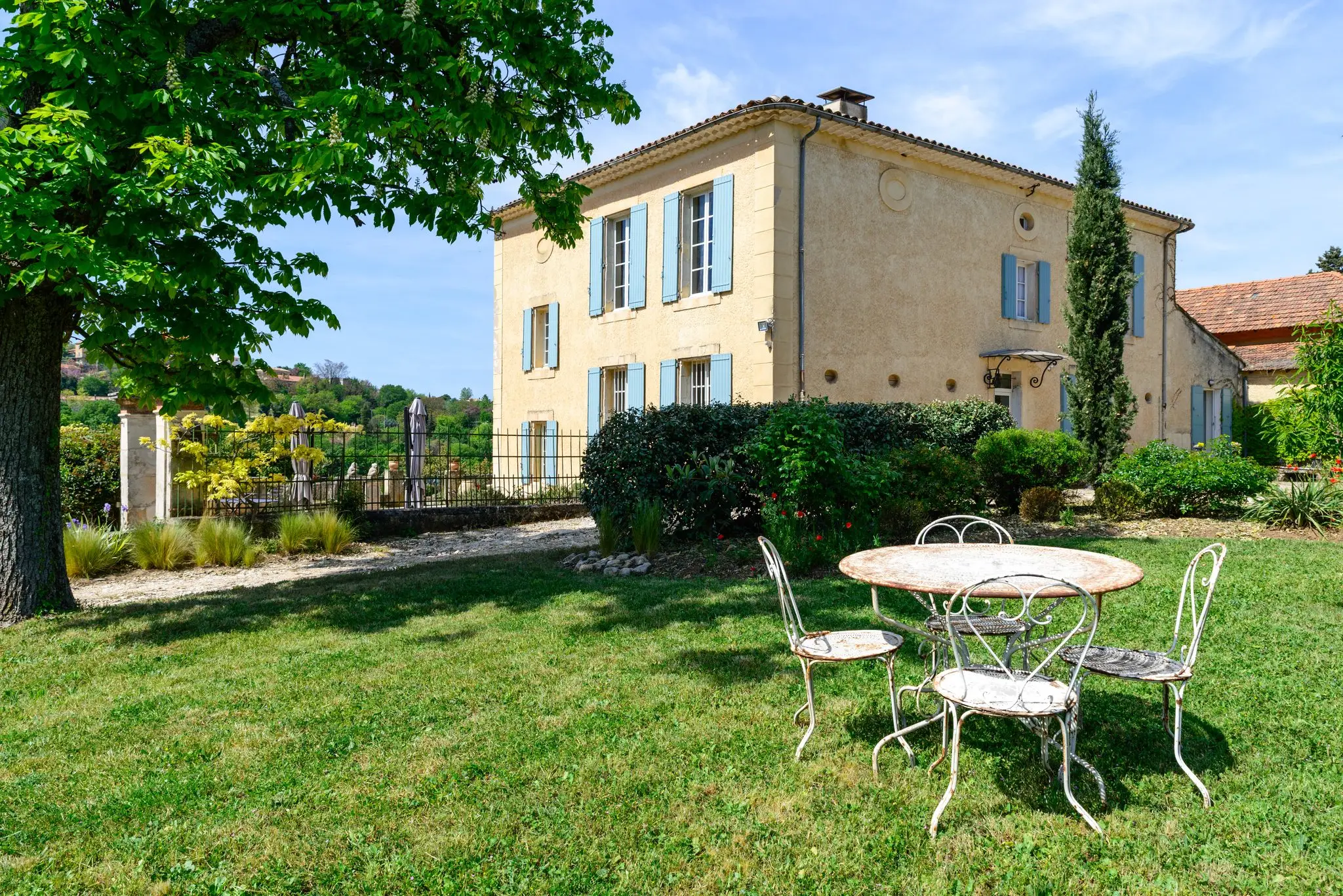 Bastide ocre aux volets bleu-gris vue depuis la pelouse, table et chaises de jardin en fer blanc à l'ombre d'un grand arbre