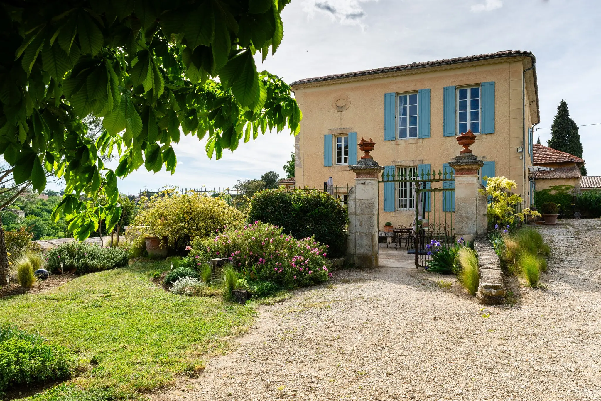 Bastide aux murs ocre et volets bleu-gris derrière un portail en fer forgé encadré de piliers en pierre et de massifs fleuris
