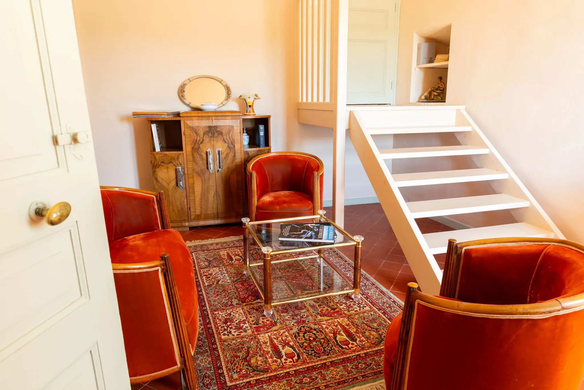 Coin salon avec fauteuils en velours orange, tapis d'Orient, meuble Art déco en ronce de noyer et escalier blanc vers une mezzanine