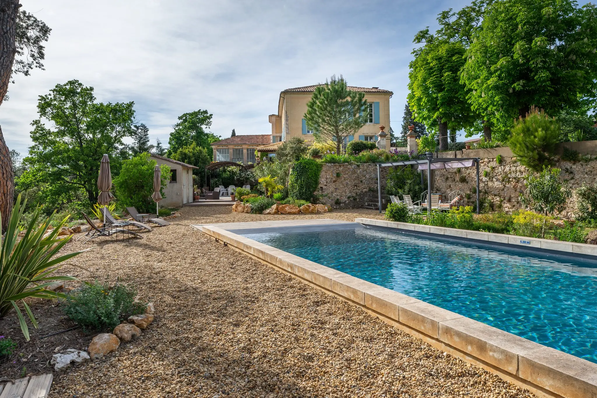 Piscine aux margelles de pierre bordée de gravier et de plantes sèches, la bastide en fond derrière les arbres