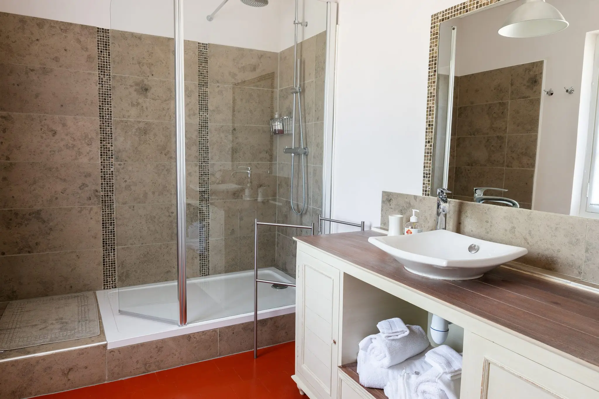Salle de bains au carrelage de pierre beige, grande douche vitrée, vasque à poser sur meuble blanc et serviettes roulées
