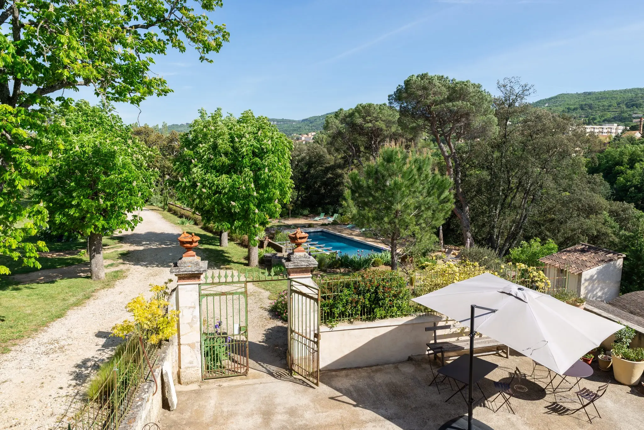 Allée, portail en pierre, terrasse ombragée et piscine au milieu des pins, collines boisées à l'horizon