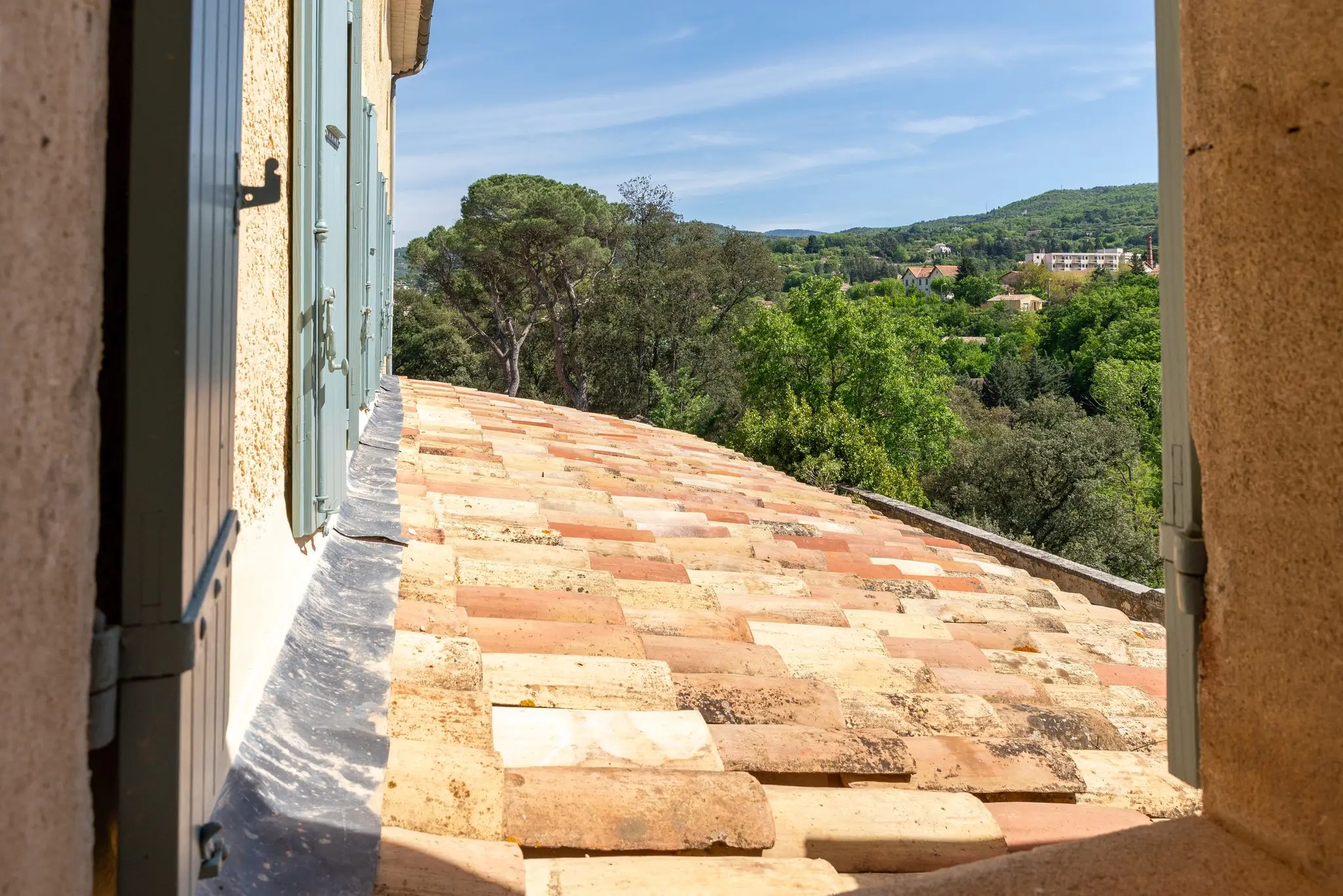 Toit de tuiles anciennes vu depuis une fenêtre aux volets bleu-gris, pins et collines boisées au loin