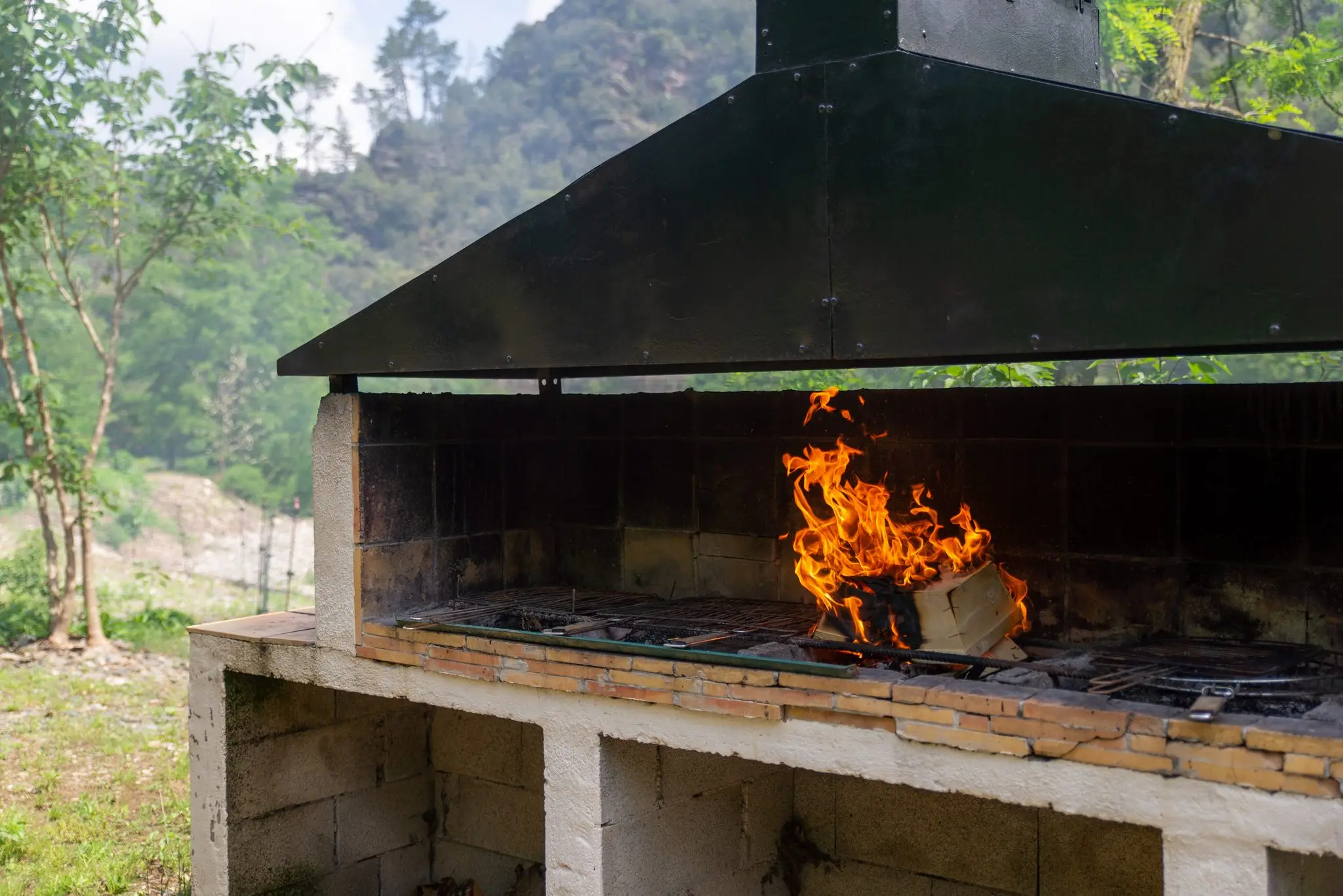 Grand barbecue maçonné sous hotte métallique, flammes vives sur la grille, végétation en arrière-plan