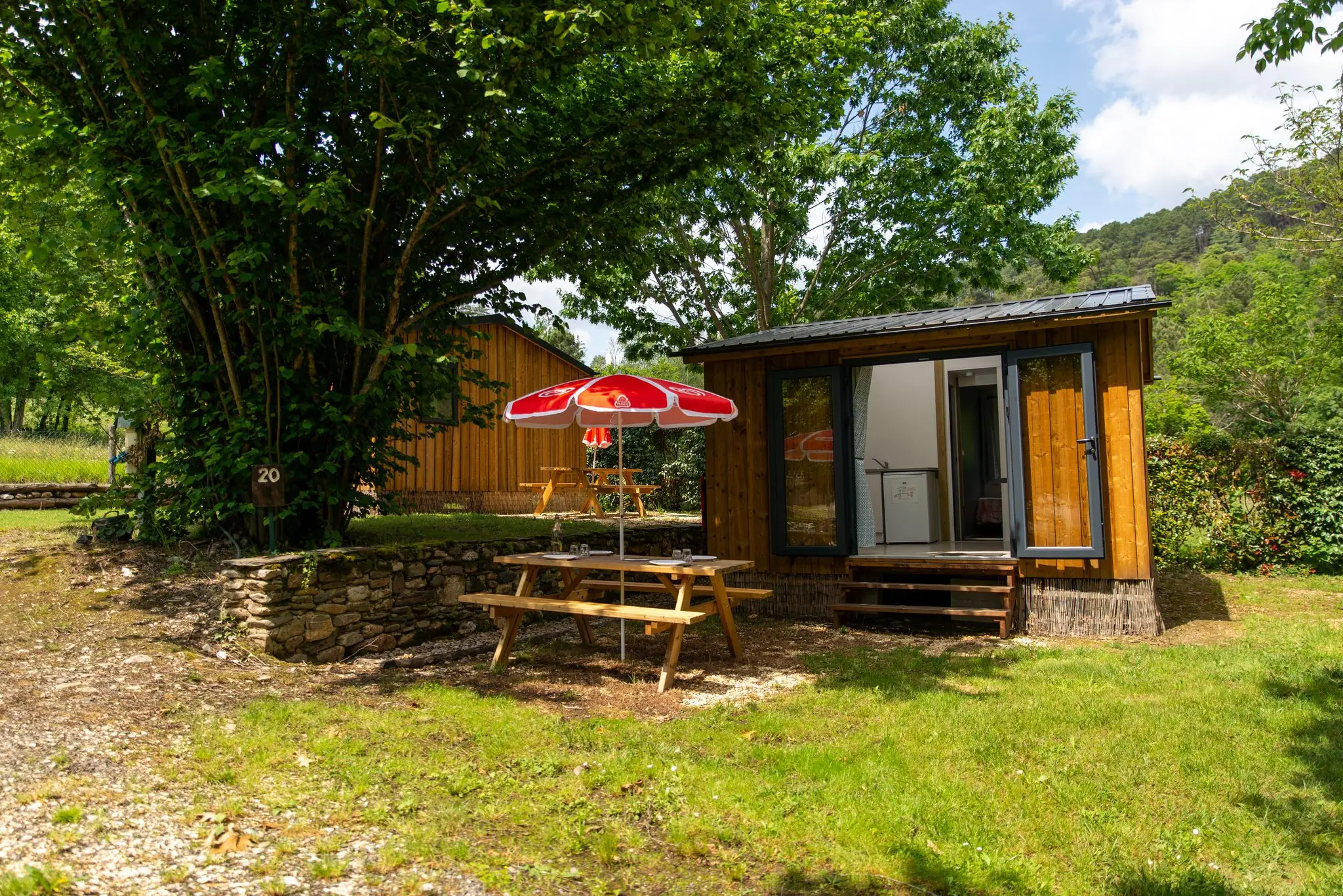 Petit chalet en bardage bois à baie vitrée, table de pique-nique et parasol rouge, à l'ombre d'un grand arbre