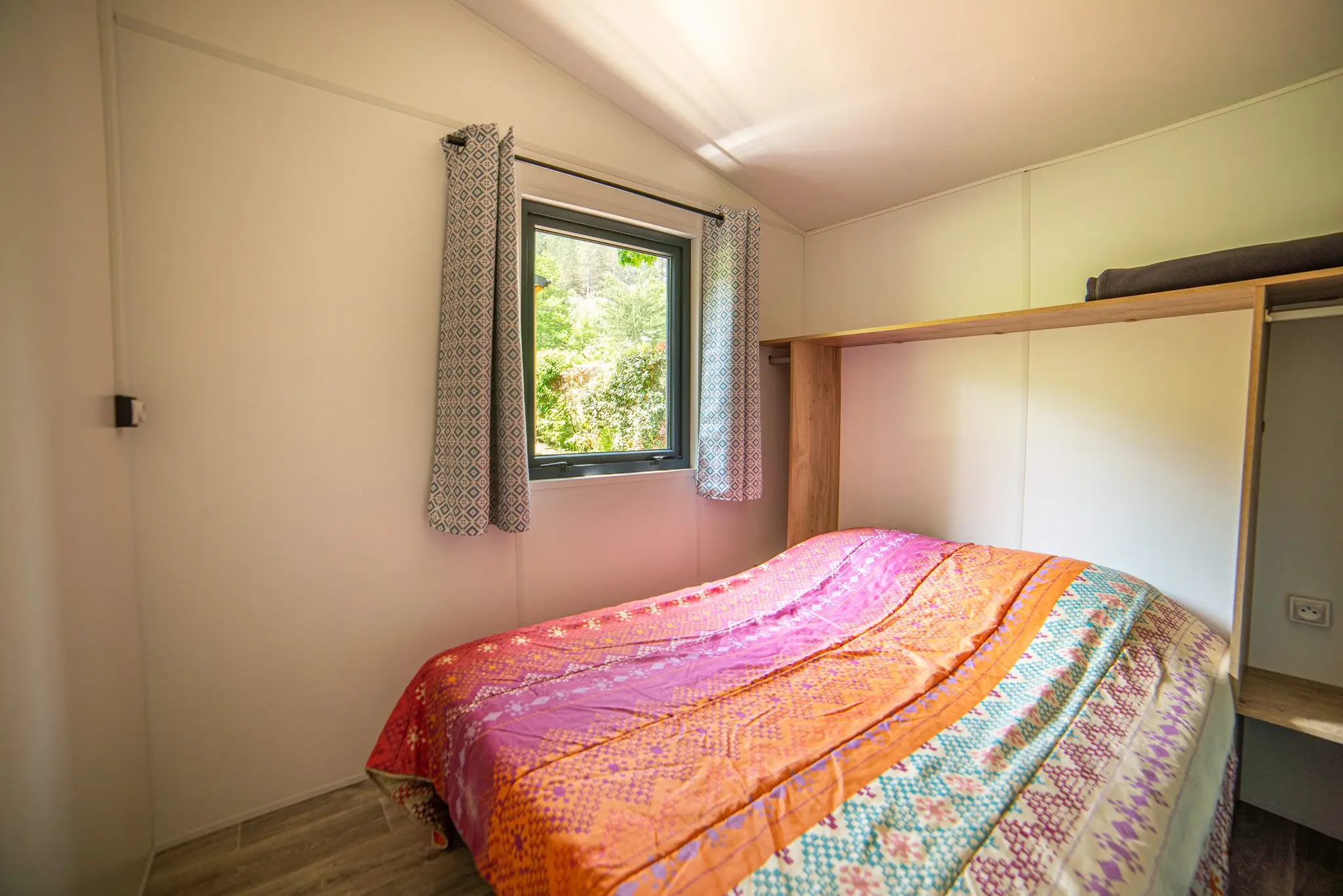 Petite chambre de chalet, lit double au couvre-lit coloré et fenêtre donnant sur la verdure