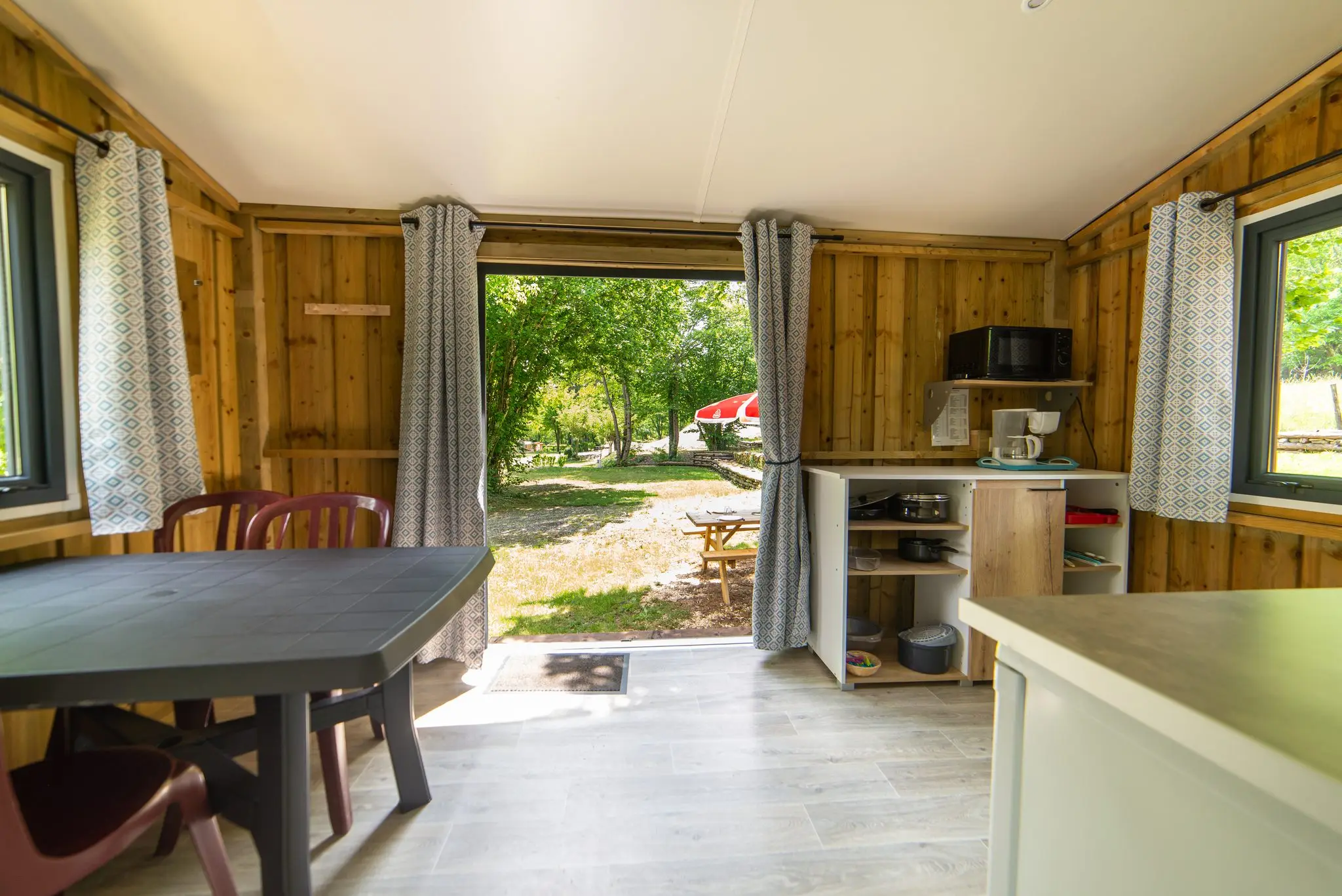 Séjour de chalet en bois avec table, kitchenette équipée et baie ouverte sur le terrain arboré
