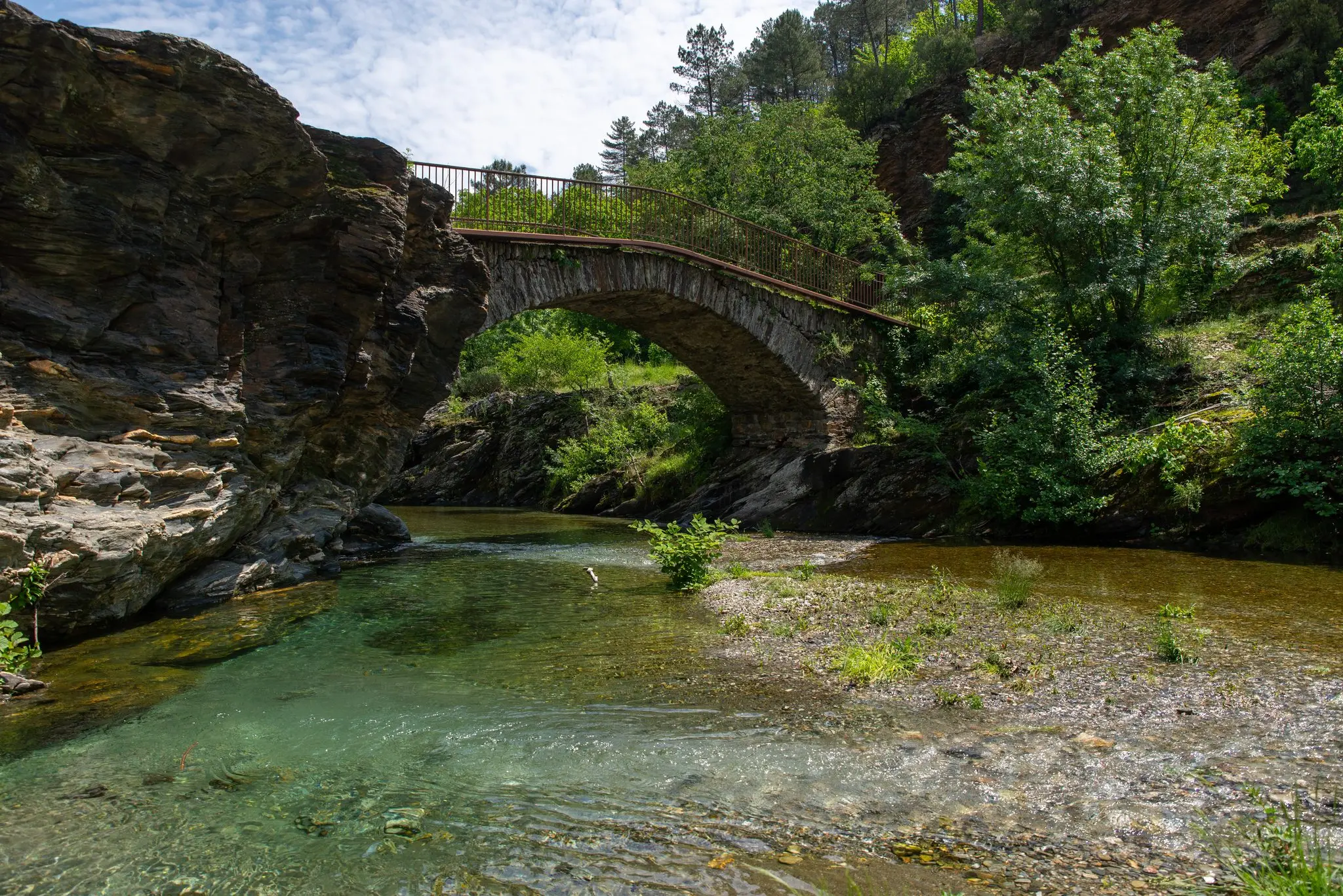 Pont ancien en arche de pierre enjambant une rivière aux eaux vertes et transparentes, entre rochers schisteux et végétation dense