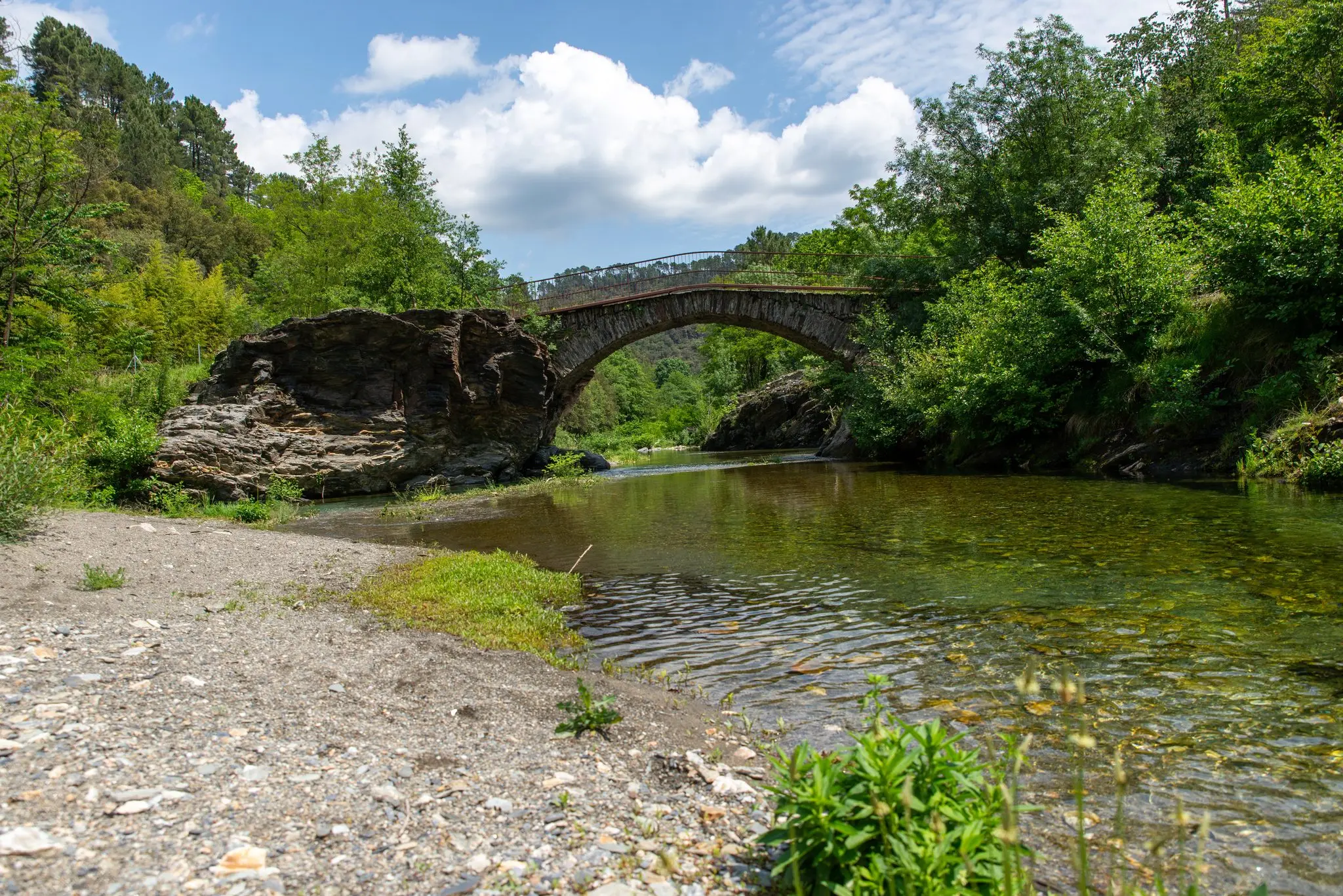 Grève de galets et eau claire d'une rivière, vieux pont de pierre en arche au loin sous un ciel nuageux