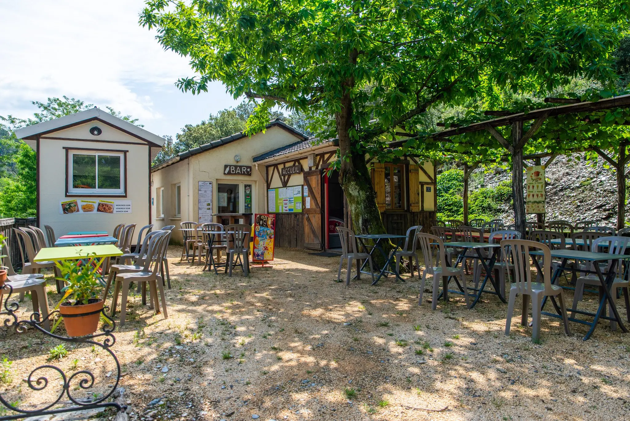 Terrasse ombragée d'un bar-snack de camping, tables et chaises de jardin sous une pergola et un grand arbre