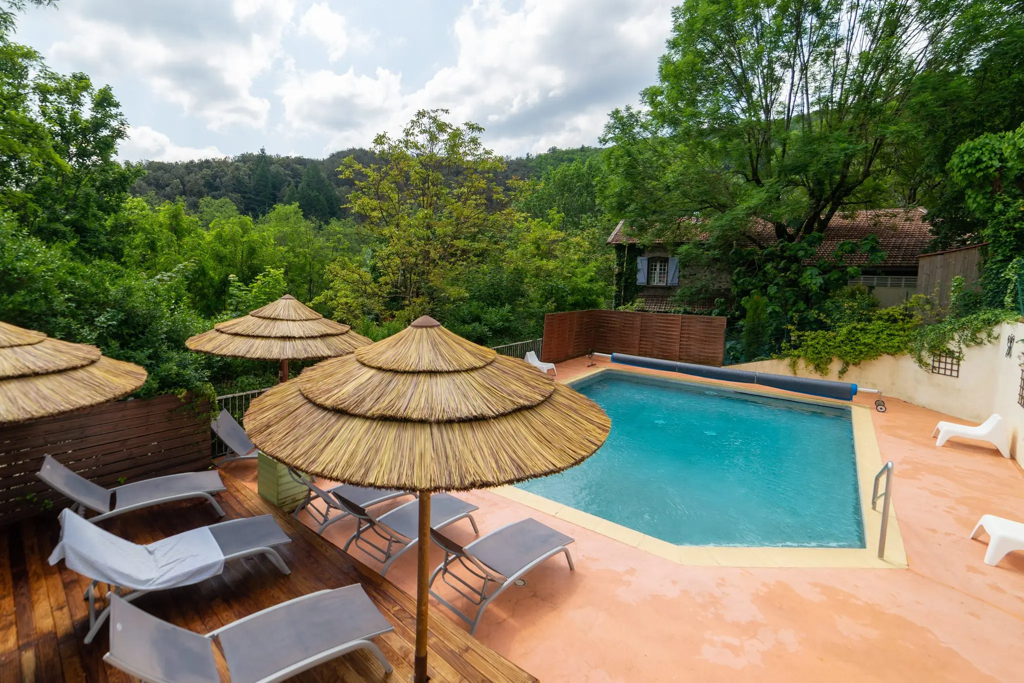Piscine de camping bordée de transats blancs et de parasols en paille, forêt en arrière-plan