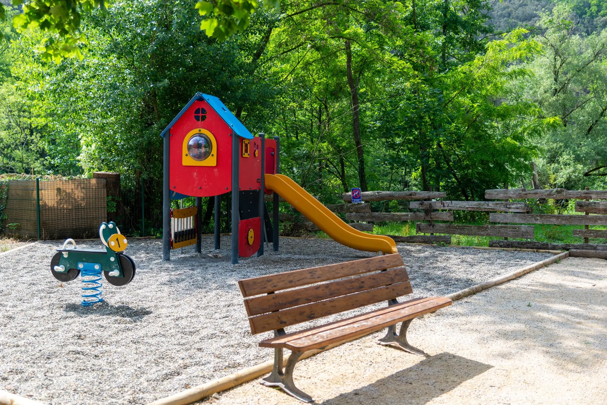 Maisonnette à toboggan jaune, jeu à ressort et banc en bois sur sol gravillonné, forêt en arrière-plan
