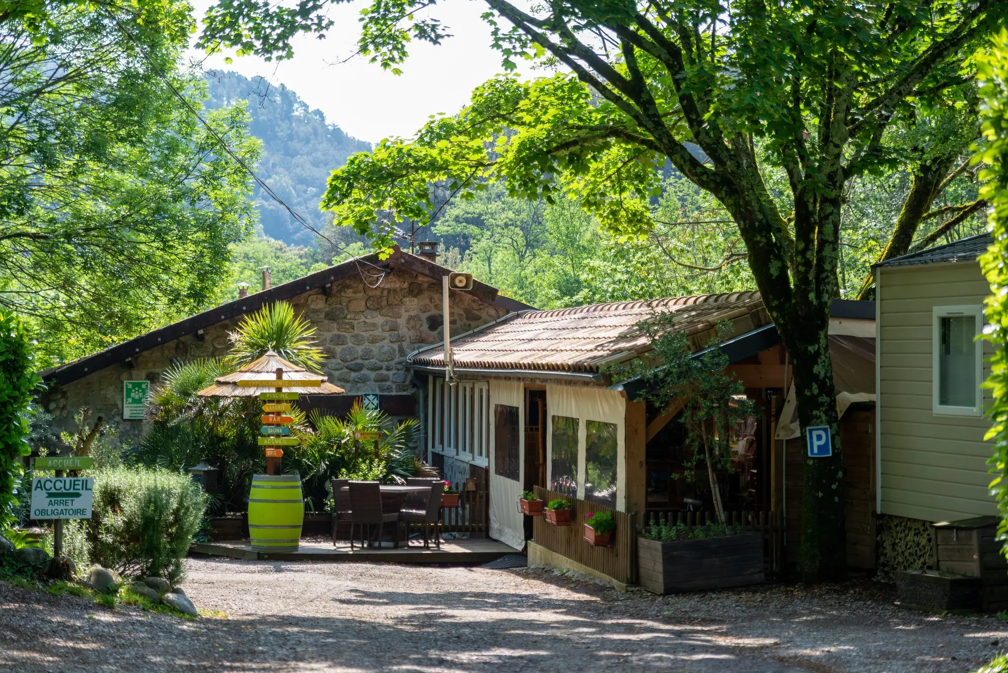 Allée gravillonnée sous les arbres menant à un bâtiment en pierre et bois avec terrasse, panneau d'accueil et tonneau fleuri