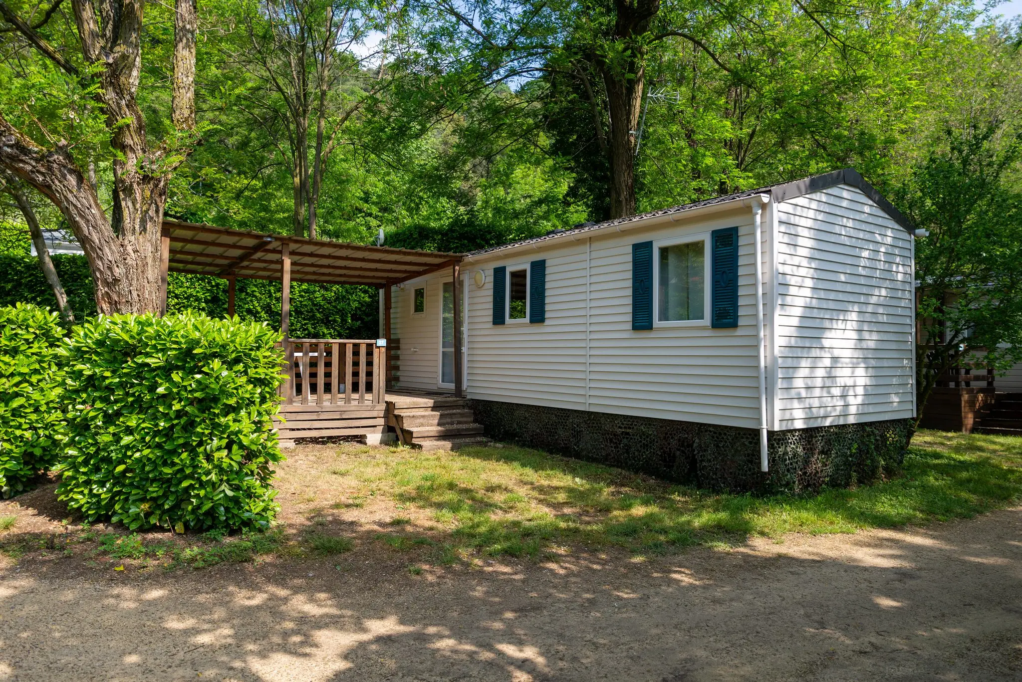 Mobil-home blanc à volets bleus, terrasse en bois couverte, haie taillée et grands arbres