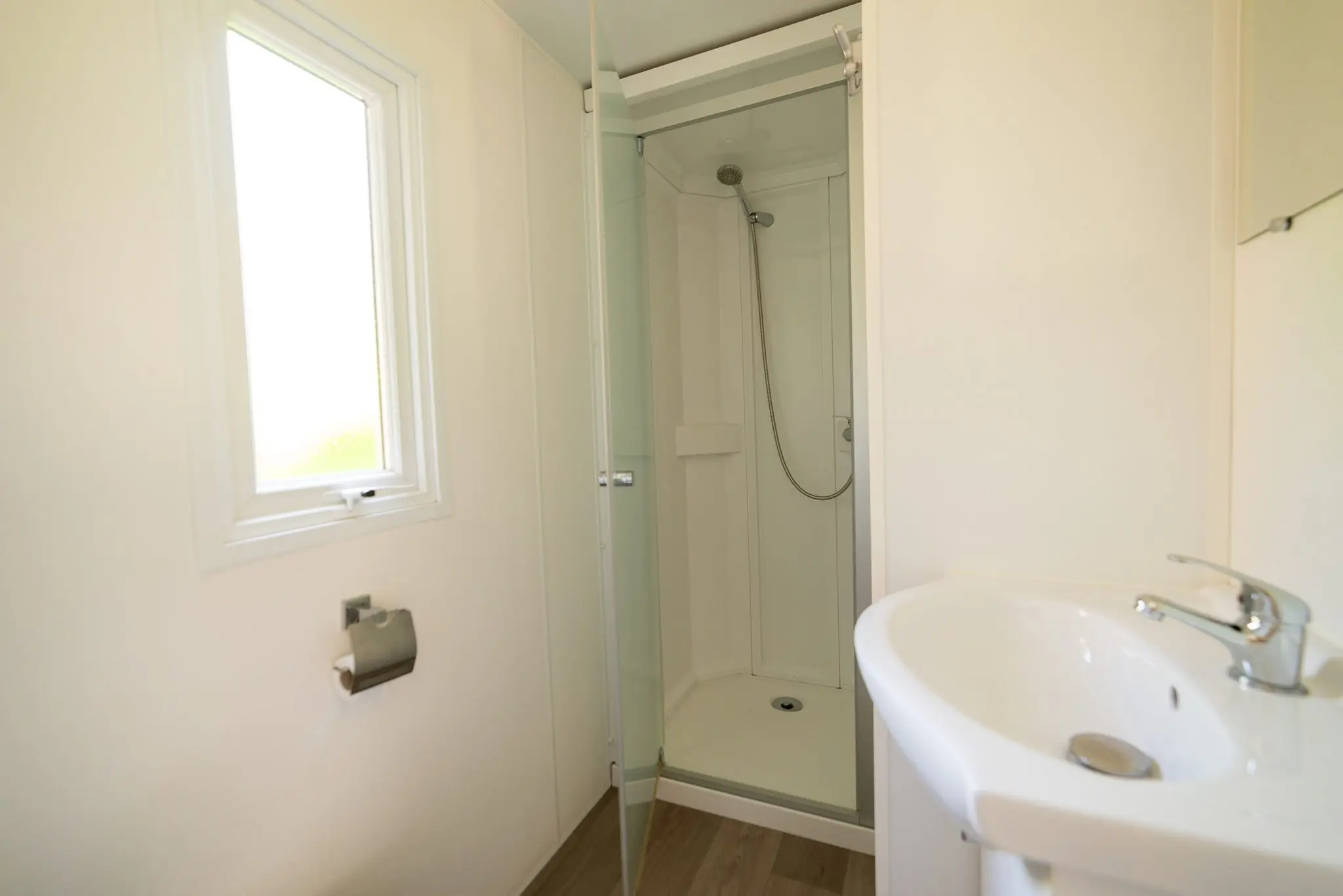 Cabine de douche vitrée, lavabo blanc et petite fenêtre dans une salle d'eau claire