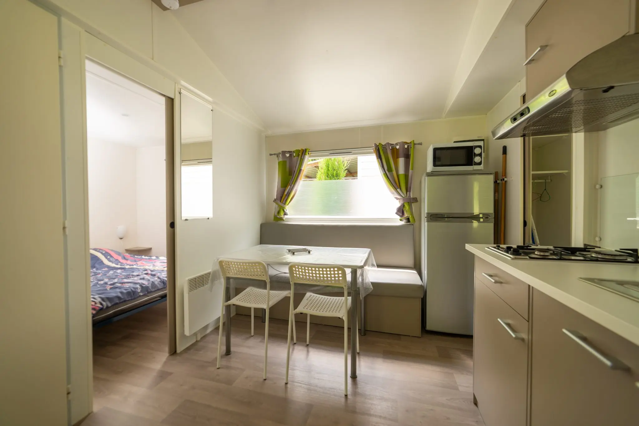Coin repas avec table et chaises, banquette, réfrigérateur, micro-ondes et plaques de cuisson, chambre visible par la porte