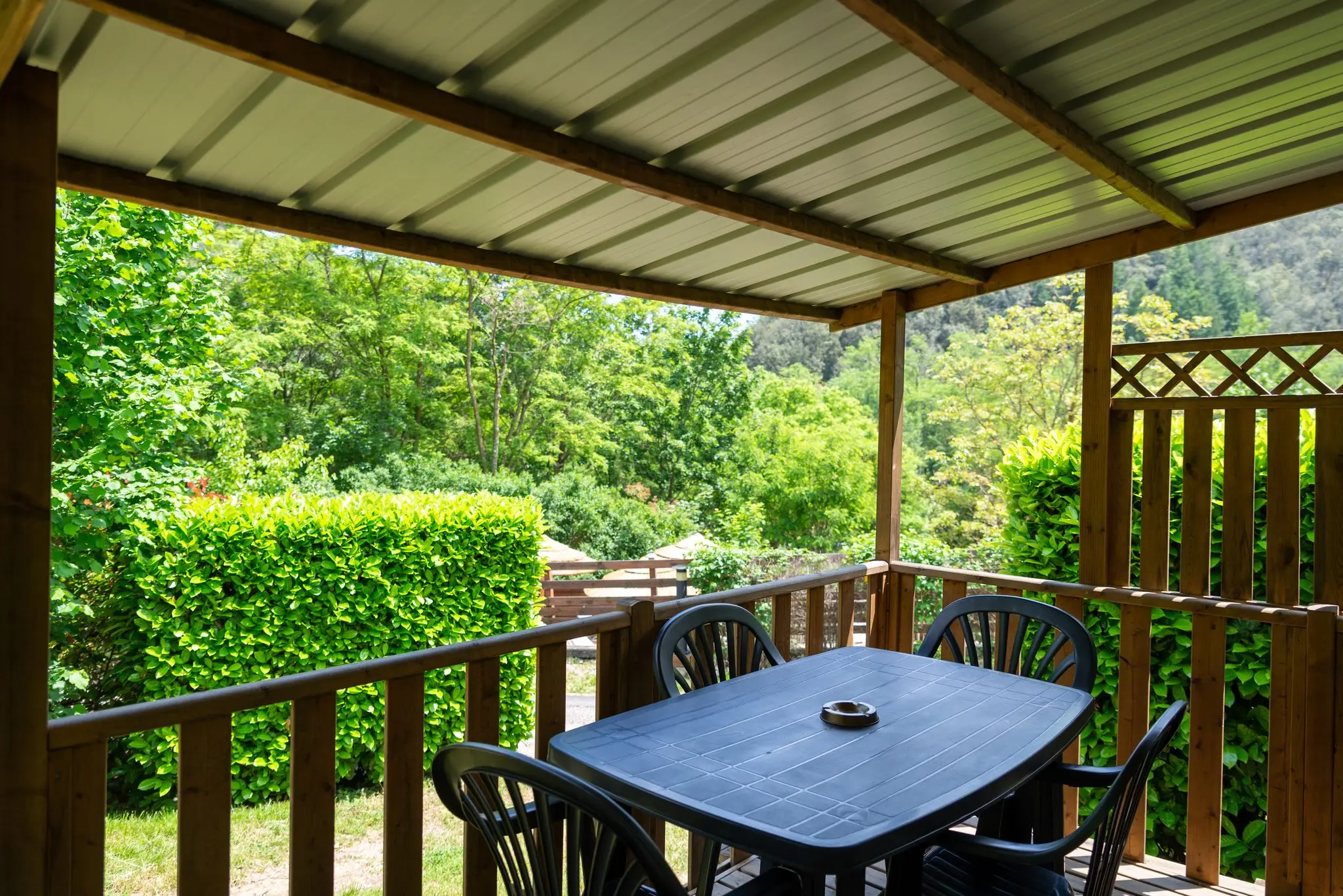 Terrasse couverte en bois avec table et chaises, haies et coteau boisé en vue