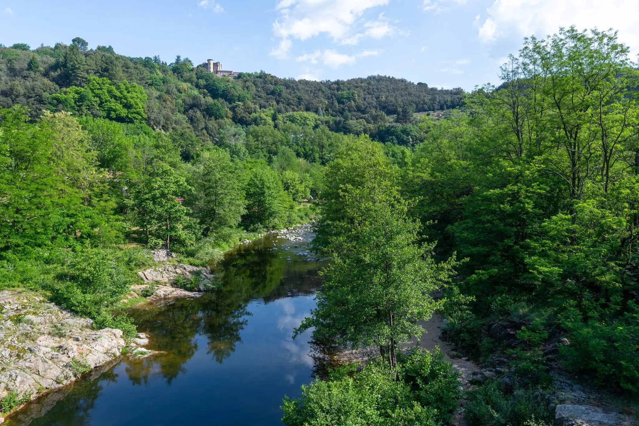 Rivière calme bordée de rochers et de forêt, collines verdoyantes et ruine perchée sous un ciel bleu, en Ardèche