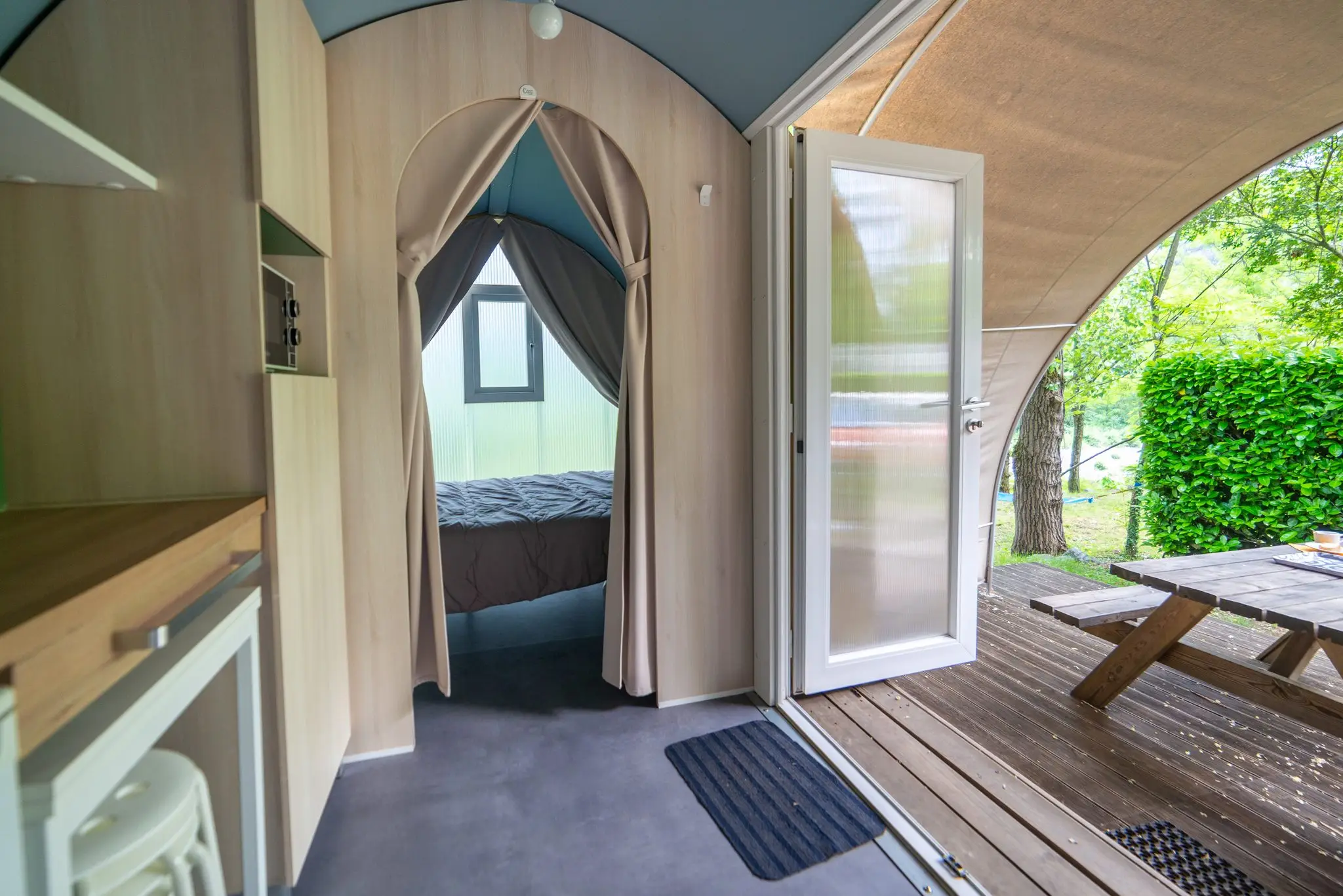 Coin cuisine en bois clair, chambre derrière un rideau en arche, porte vitrée ouverte sur la terrasse et sa table
