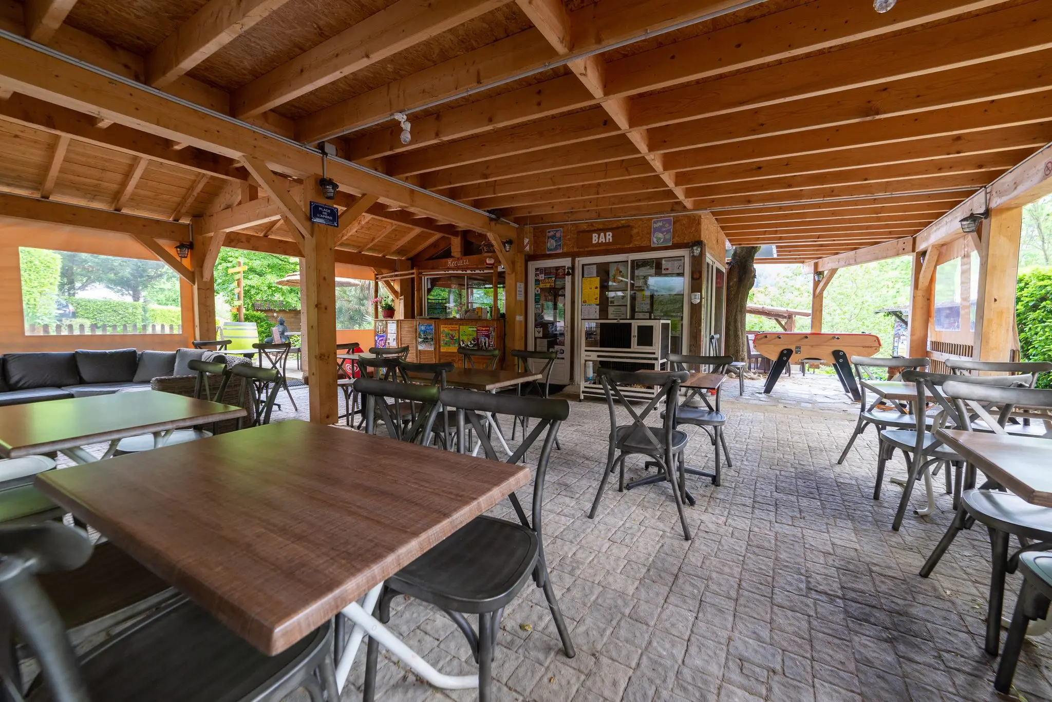 Grande terrasse sous charpente apparente, tables en bois, chaises bistrot, salon de jardin et comptoir du bar en fond