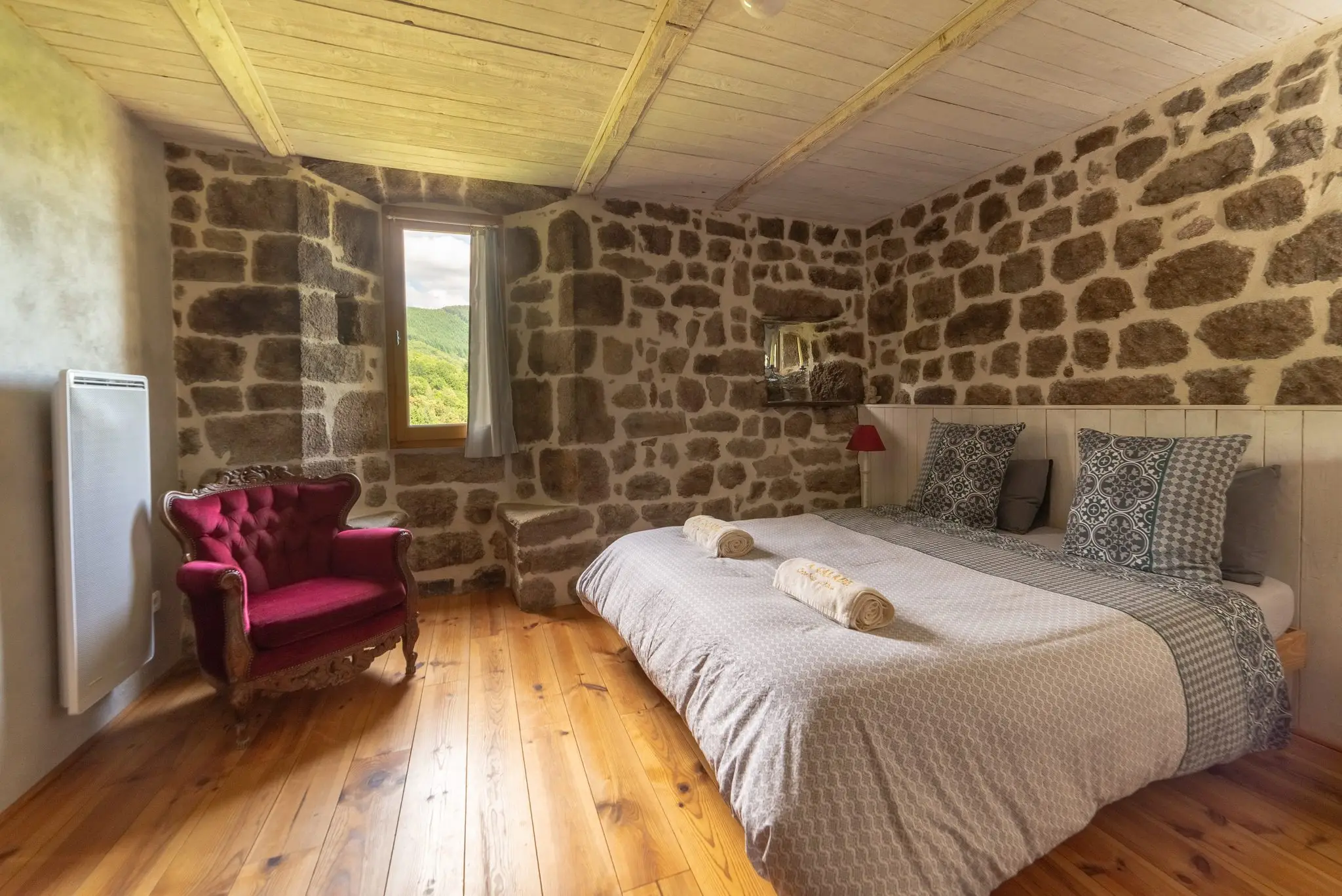 Chambre au parquet de pin, murs en pierre apparente, lit double aux serviettes roulées, fauteuil crapaud rouge et petite fenêtre sur la verdure