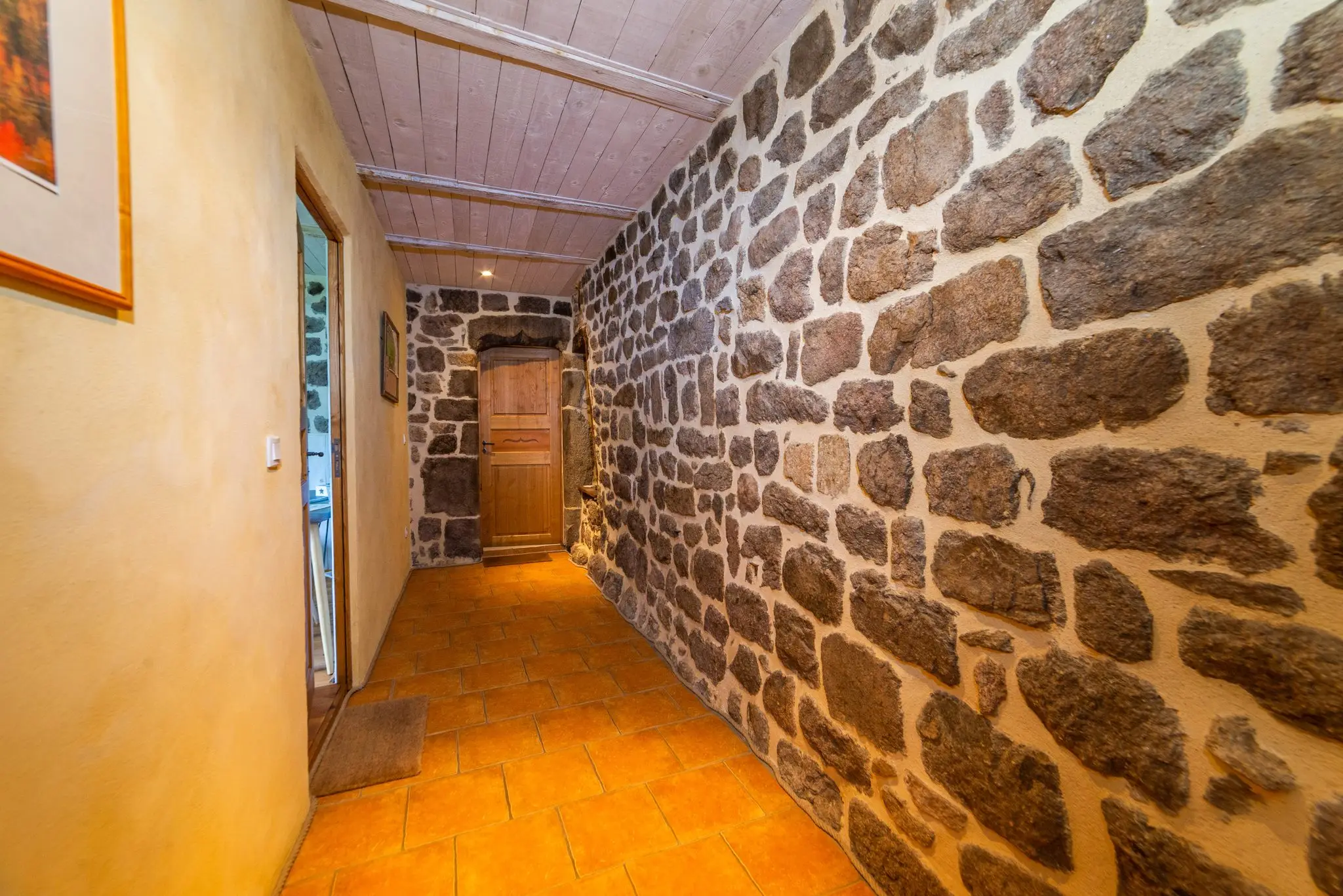 Couloir étroit au sol en tomettes, longé d'un mur en moellons de pierre, plafond en lambris blanchi et porte en bois au fond