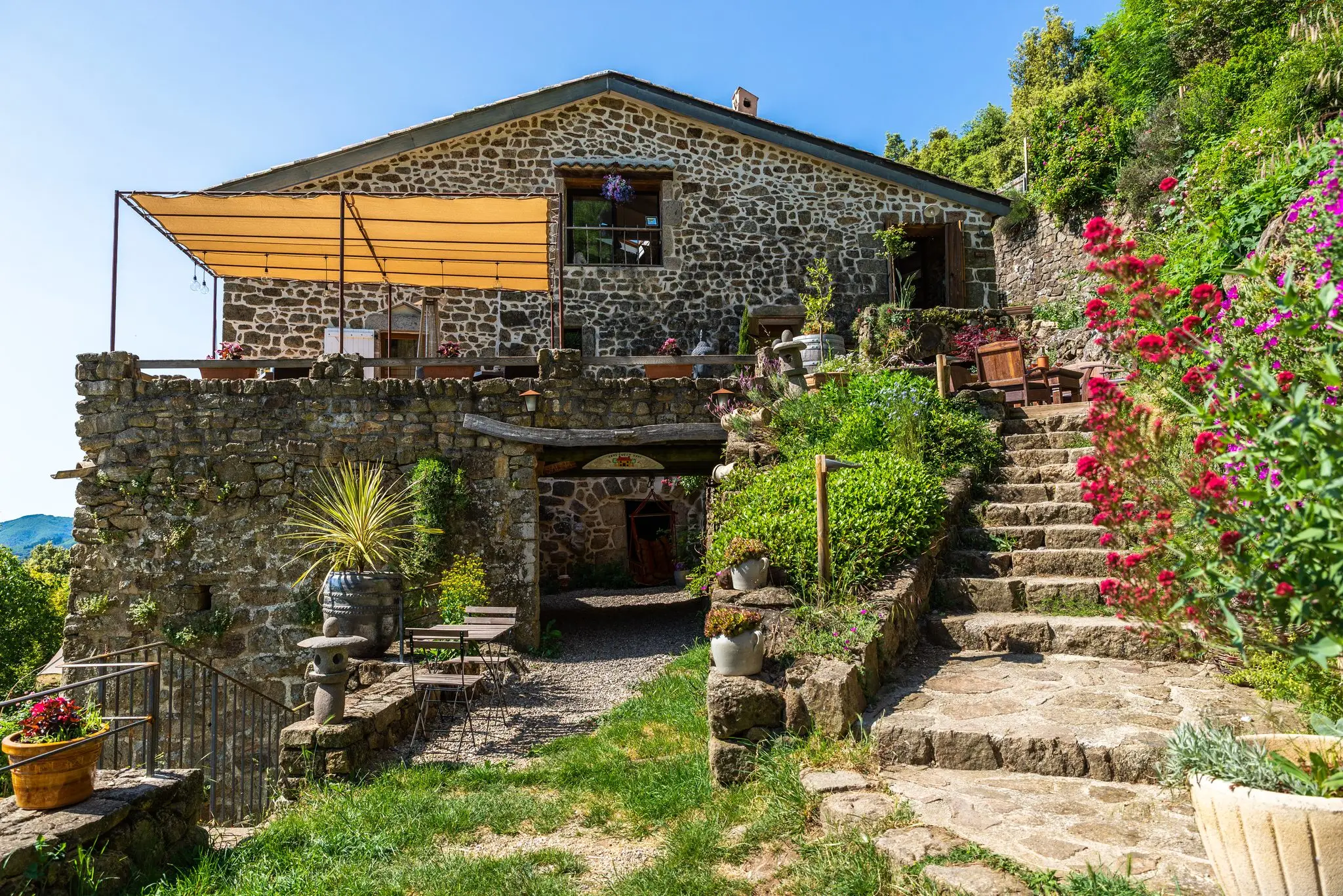 Façade d'une maison en pierre de pays avec terrasse sous toile tendue, escalier en pierre bordé de valérianes et de lampions, pelouse au premier plan