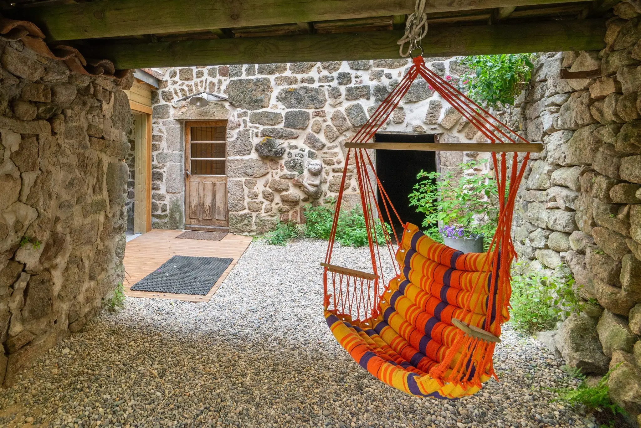 Fauteuil hamac rayé orange suspendu sous un auvent, cour gravillonnée entourée de murs en pierre et porte en bois