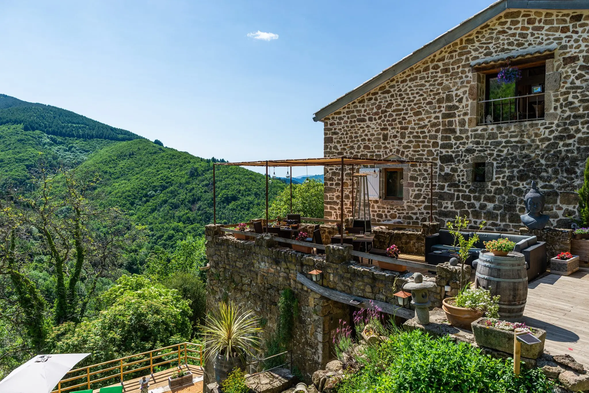 Pignon en pierre d'une maison d'hôtes dominant une terrasse sous pergola, tonneau et pots fleuris, versants boisés de la vallée en arrière-plan