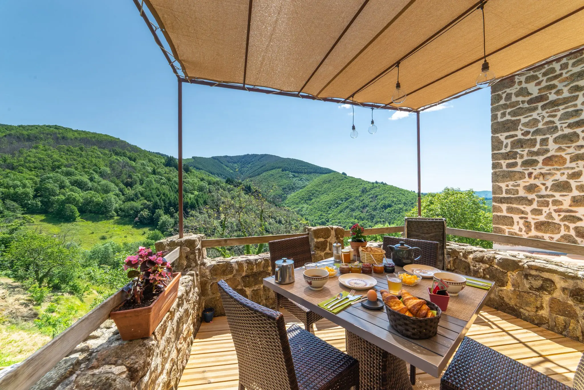 Table dressée pour le petit-déjeuner avec croissants, confitures et jus, chaises tressées, muret de pierre fleuri et vallée verdoyante sous un ciel bleu