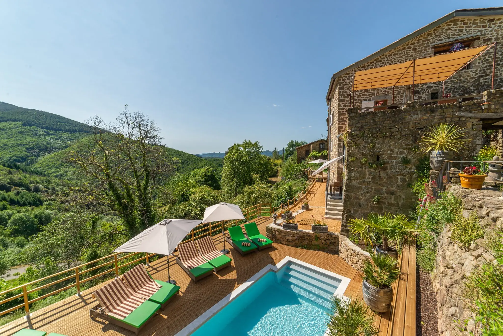 Piscine sur deck en bois et transats verts au pied d'une maison en pierre, face à la vallée boisée