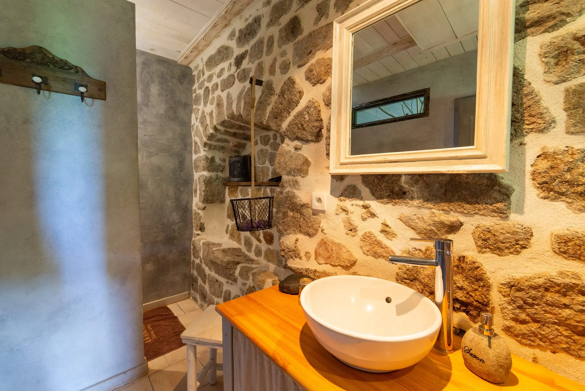 Vasque blanche sur plan en bois clair, mur en pierre apparente, miroir à cadre blanc et niche voûtée