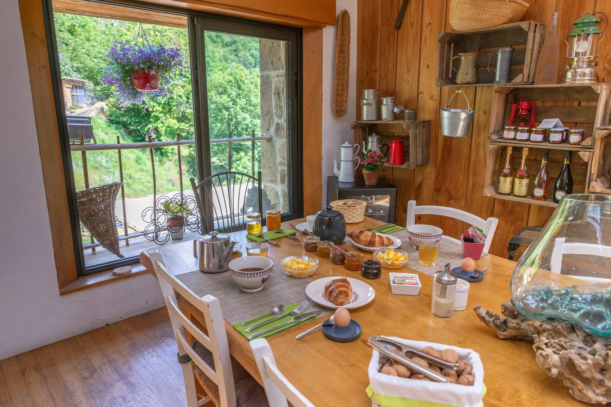 Table en bois dressée pour le petit-déjeuner, étagères en caisses de bois garnies de confitures et de bouteilles, baie vitrée sur un balcon fleuri et la verdure