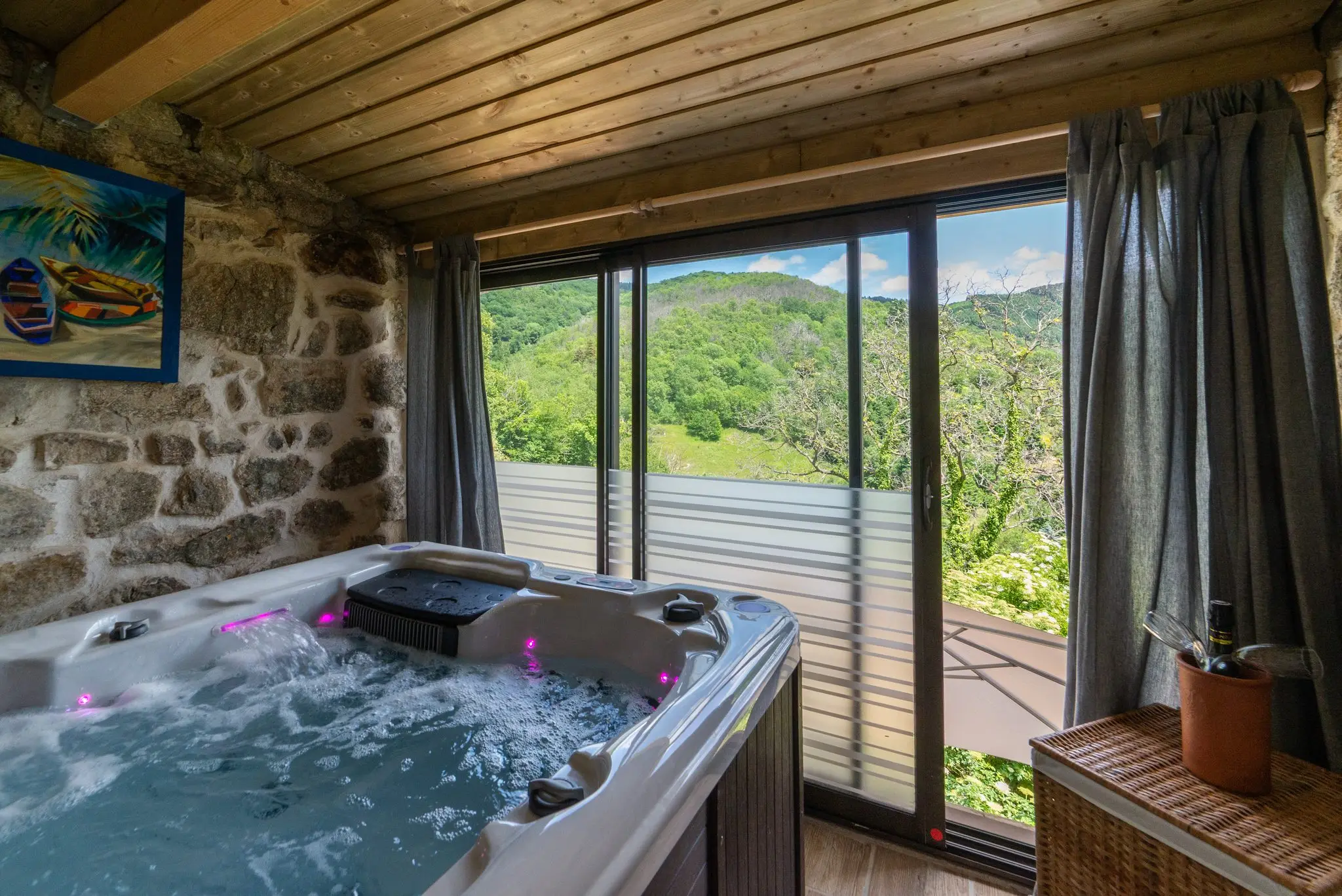 Bain à remous en marche sous un plafond en bois, mur en pierre et grande baie vitrée ouvrant sur les collines boisées