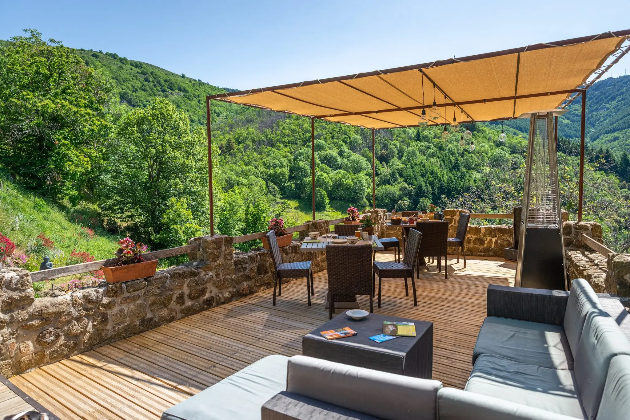 Grande terrasse en lames de bois avec salon extérieur, tables de petit-déjeuner sous une pergola en toile et muret en pierre ouvrant sur les collines boisées