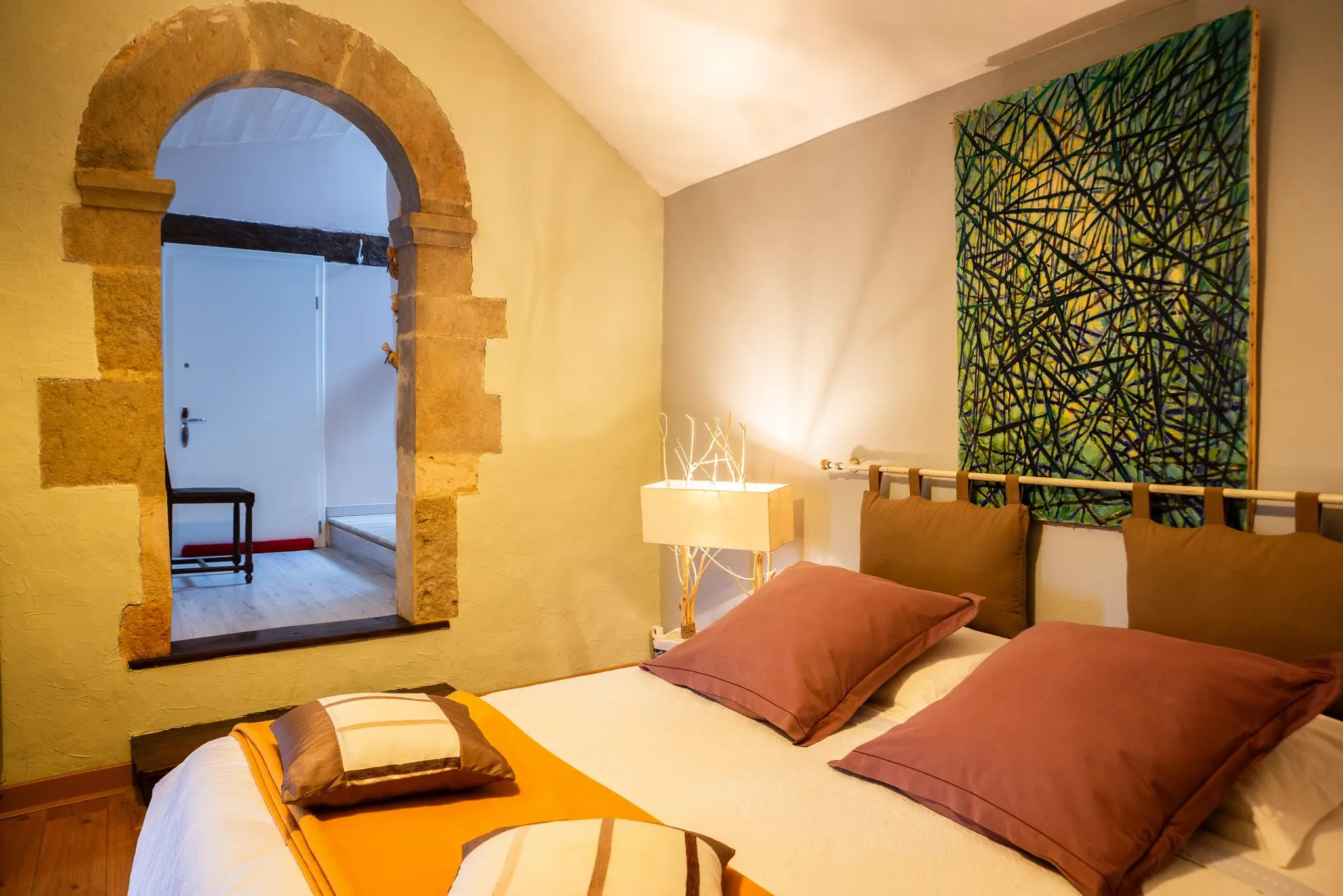 Chambre aux tons orangés, lit double aux coussins vieux rose, grande toile abstraite verte et baie intérieure en arc de pierre