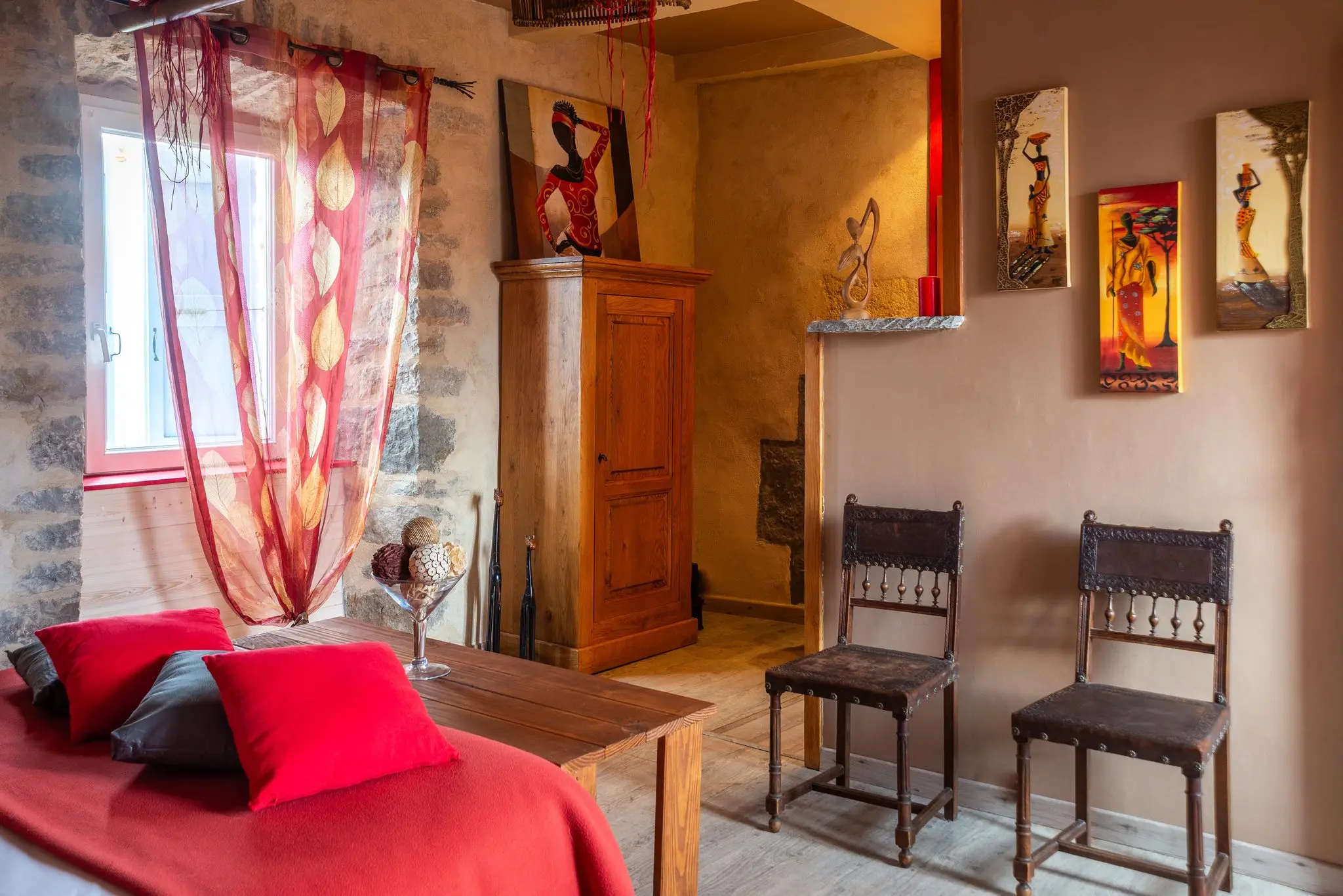 Fenêtre voilée de rouge, armoire en bois, deux chaises anciennes en cuir et tableaux de style africain dans une chambre aux murs ocre