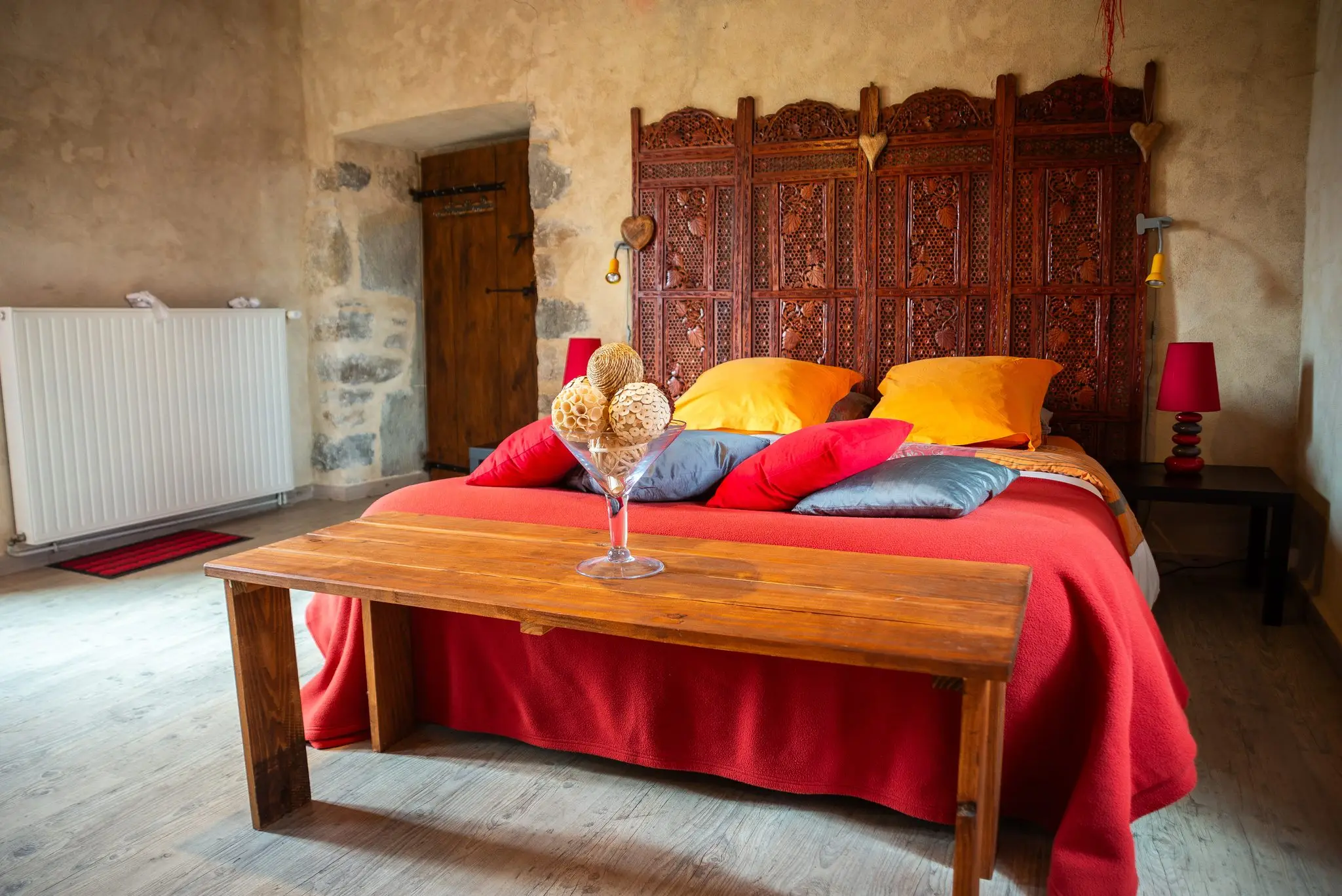 Chambre aux murs en pierre enduite, grand lit à tête de lit en bois sculpté, couvre-lit rouge, coussins orangés et banc en bois brut