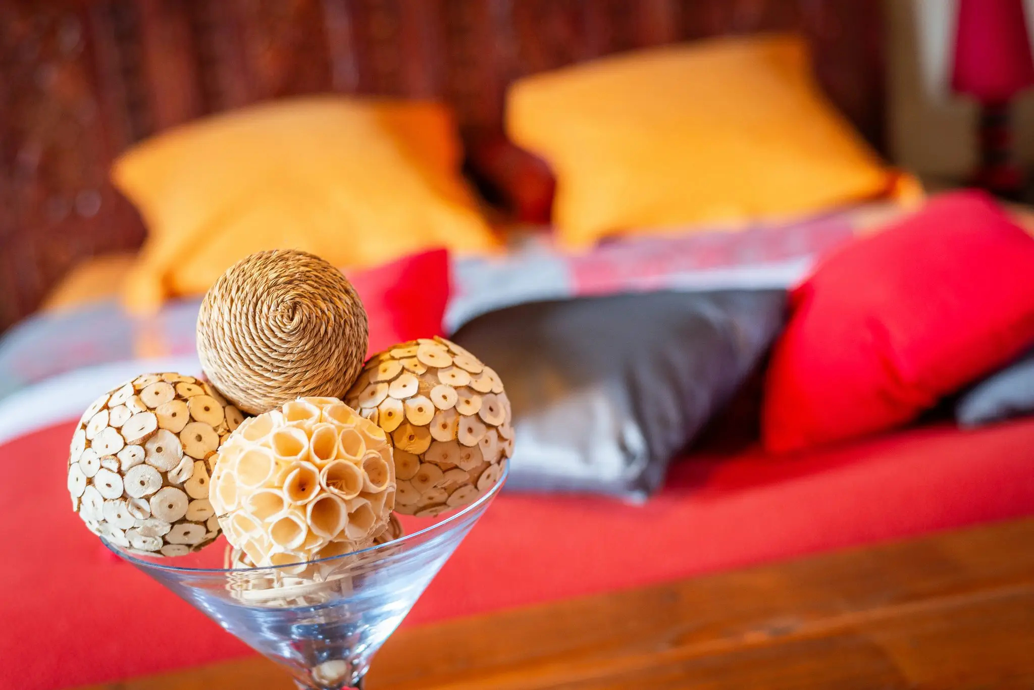 Coupe en verre garnie de boules en fibres naturelles, devant un lit au couvre-lit rouge et aux coussins orange