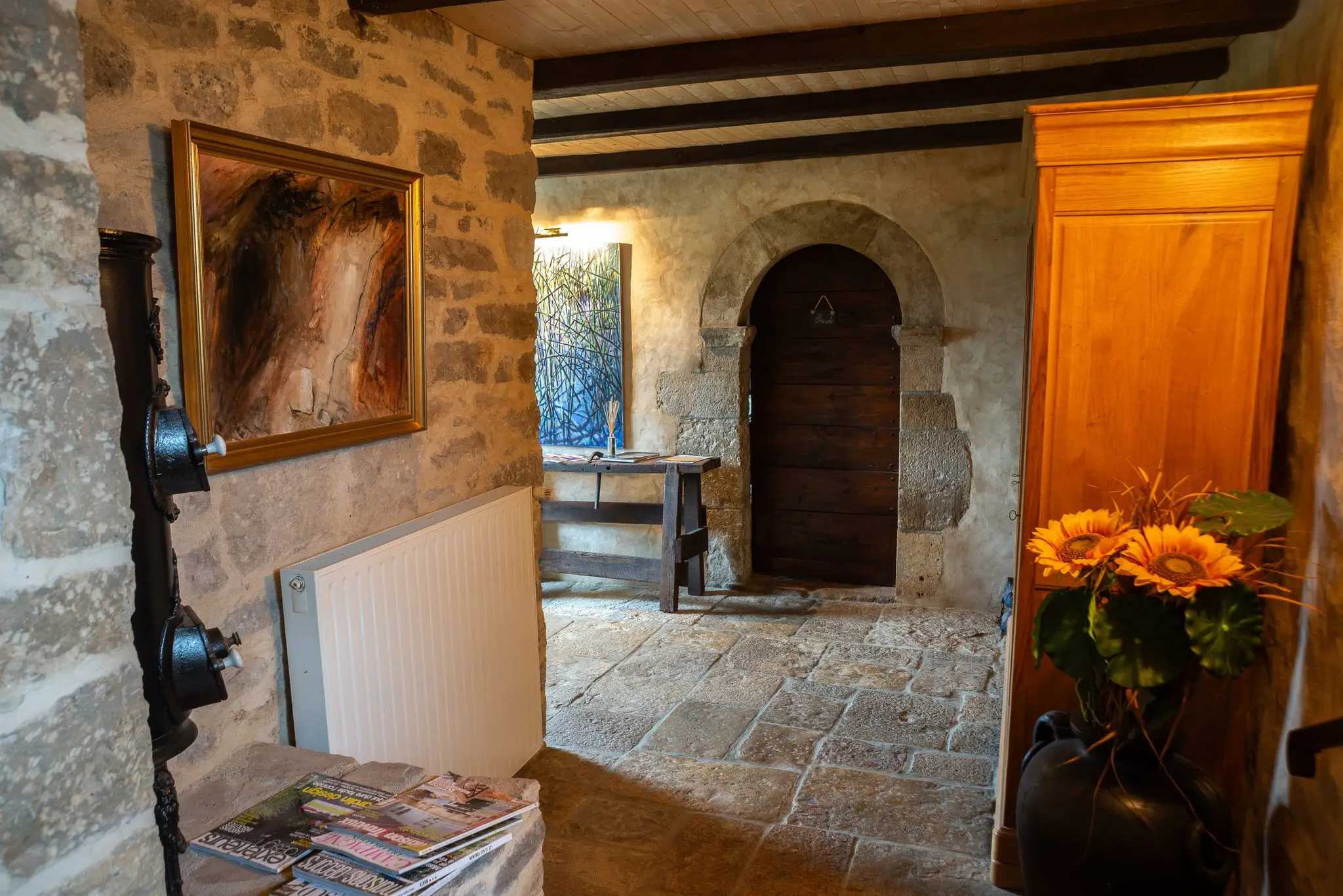Couloir d'entrée aux murs en pierre et dallage ancien, porte en bois sous arc de pierre, armoire en bois clair et bouquet de tournesols