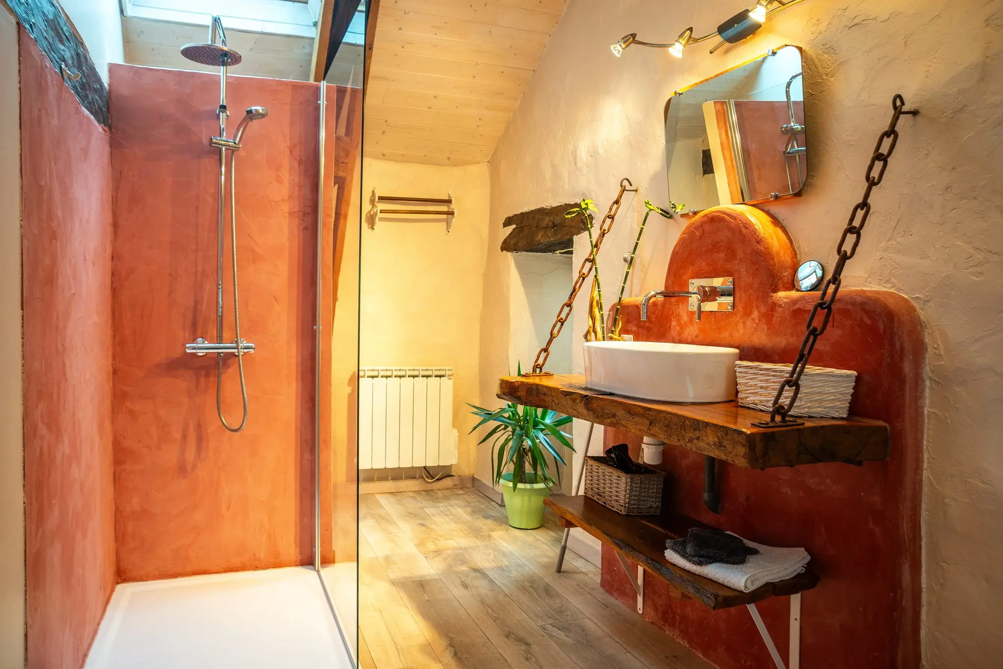 Salle d'eau sous plafond lambrissé, douche aux murs enduits couleur terre cuite, vasque sur plateau de bois brut suspendu par des chaînes