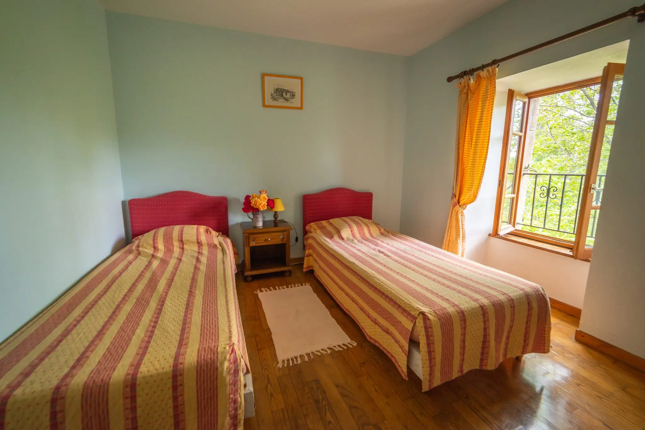 Chambre aux murs bleu pâle, deux lits simples à couvre-lits rayés, parquet et fenêtre ouverte sur les arbres