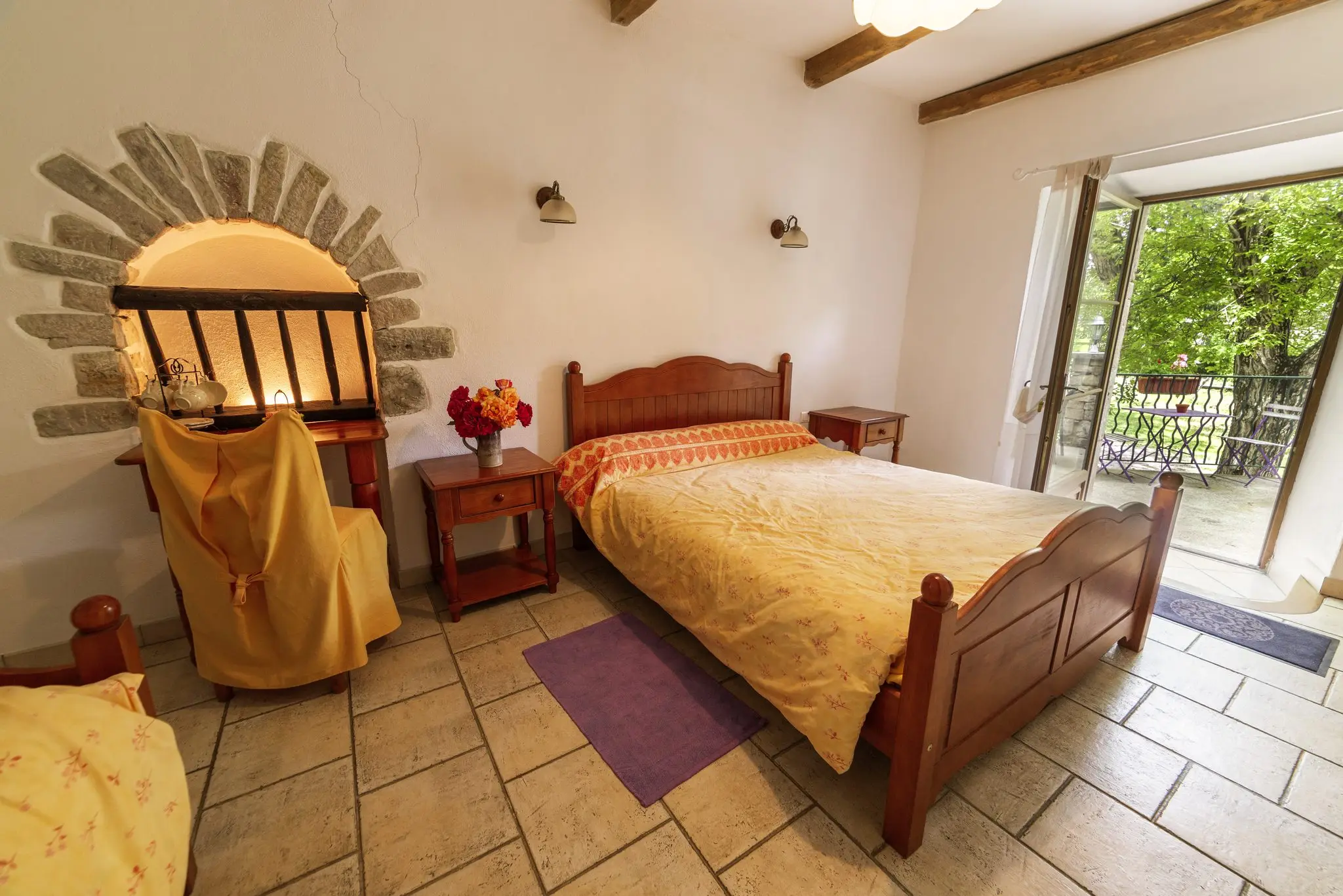 Chambre au carrelage clair, lit en bois et couvre-lit jaune, niche en arc de pierre et porte-fenêtre ouverte sur un balcon