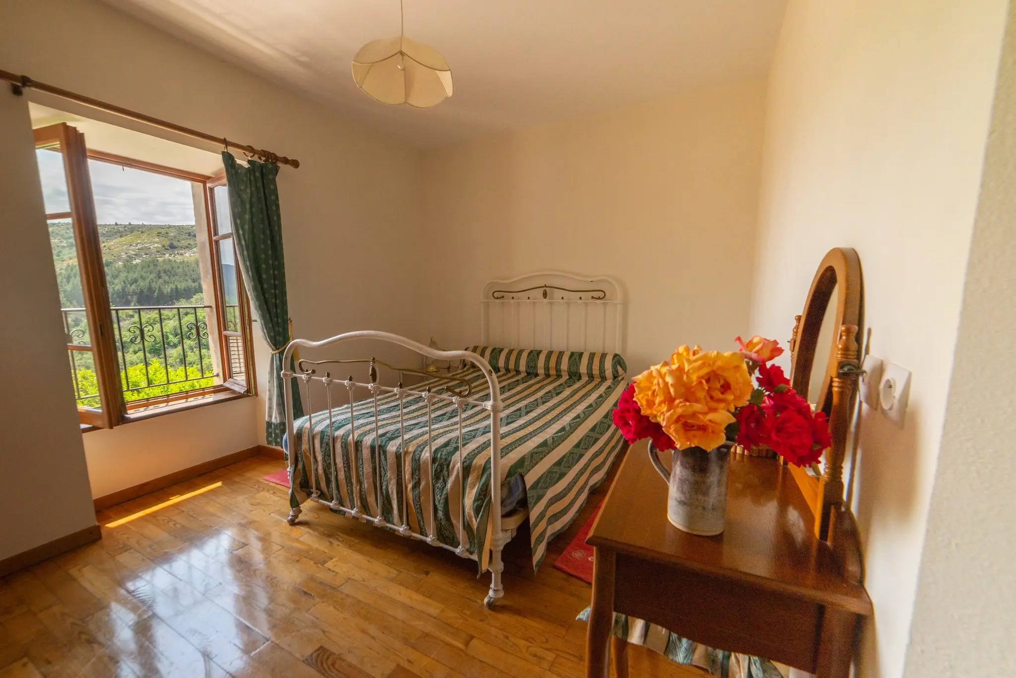 Chambre au parquet ciré, lit en fer forgé blanc, coiffeuse fleurie et fenêtre ouverte sur les crêtes boisées