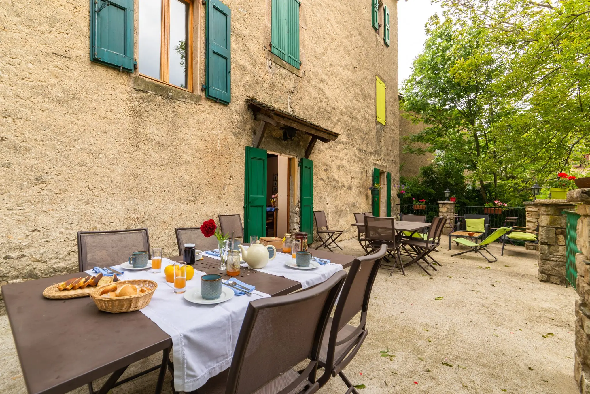 Cour gravillonnée au pied d'un mur en pierre aux volets verts, tables du petit-déjeuner dressées à l'ombre des arbres