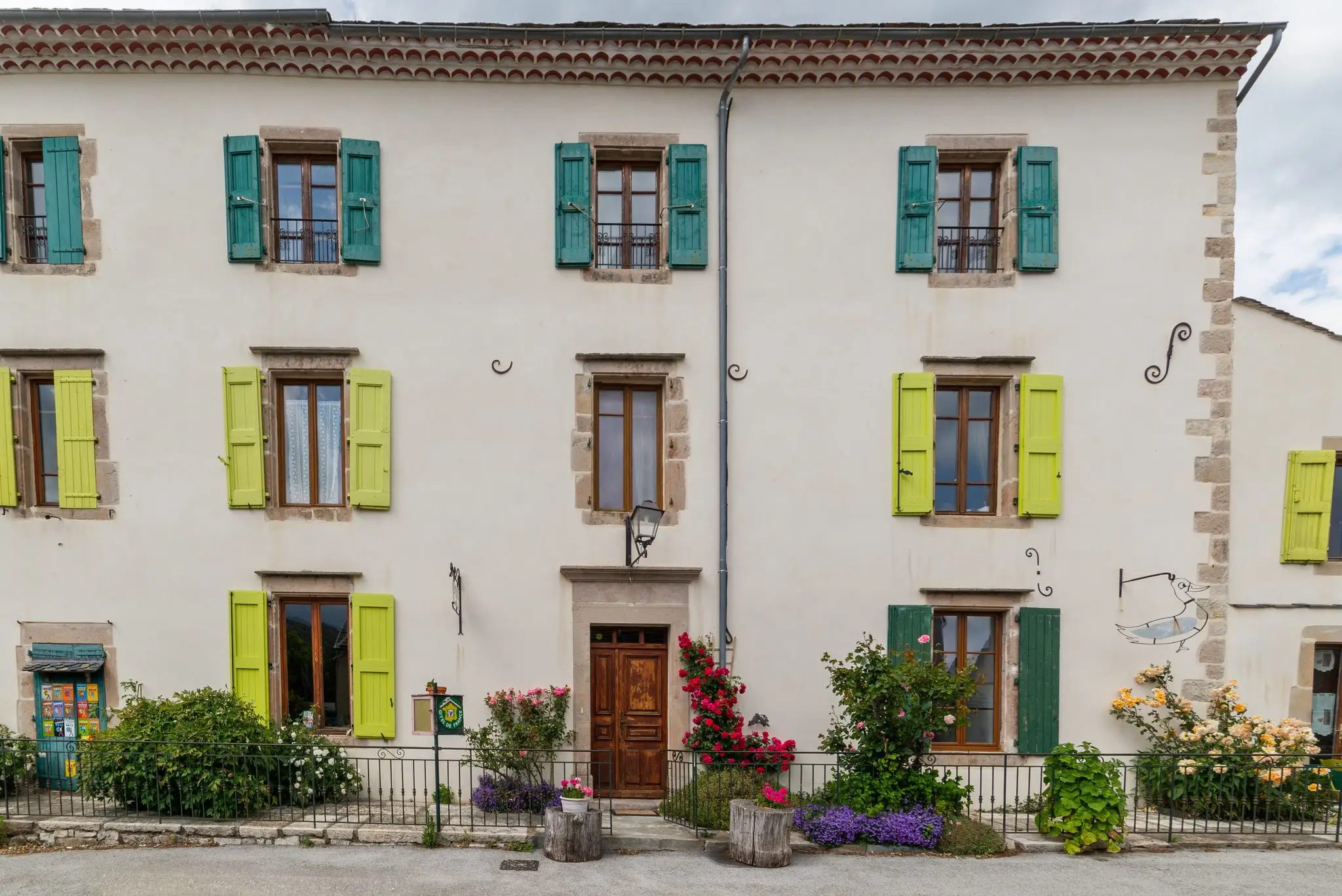 Façade blanche de maison de village à trois niveaux, volets vert anis et vert sapin, encadrements de pierre et rosiers en fleurs au pied du mur