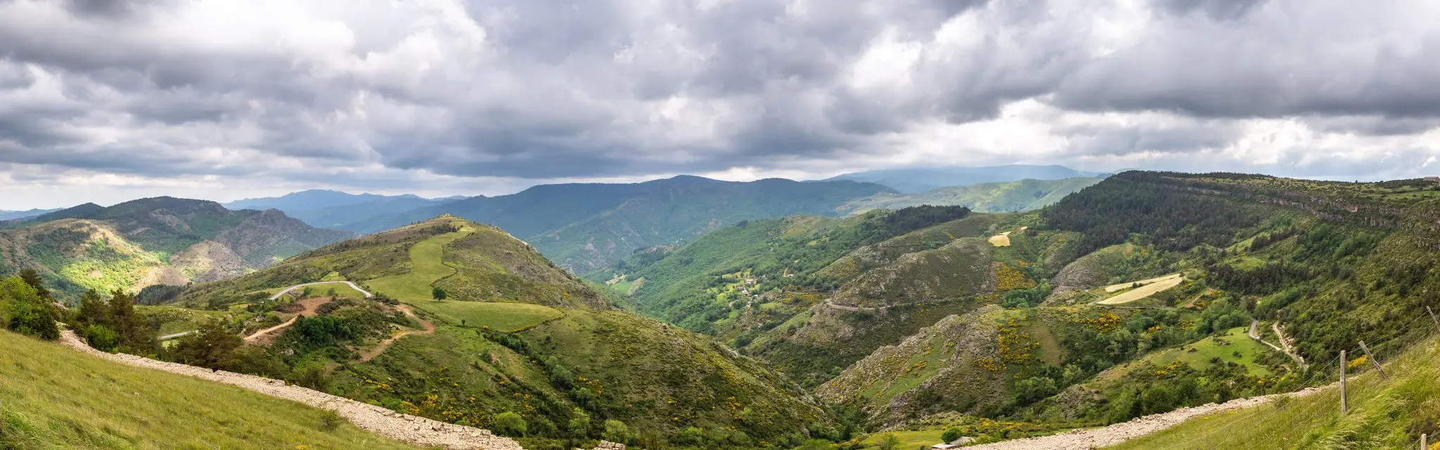 Panorama sur des vallées et crêtes verdoyantes, chemin de terre en contrebas et ciel chargé de nuages