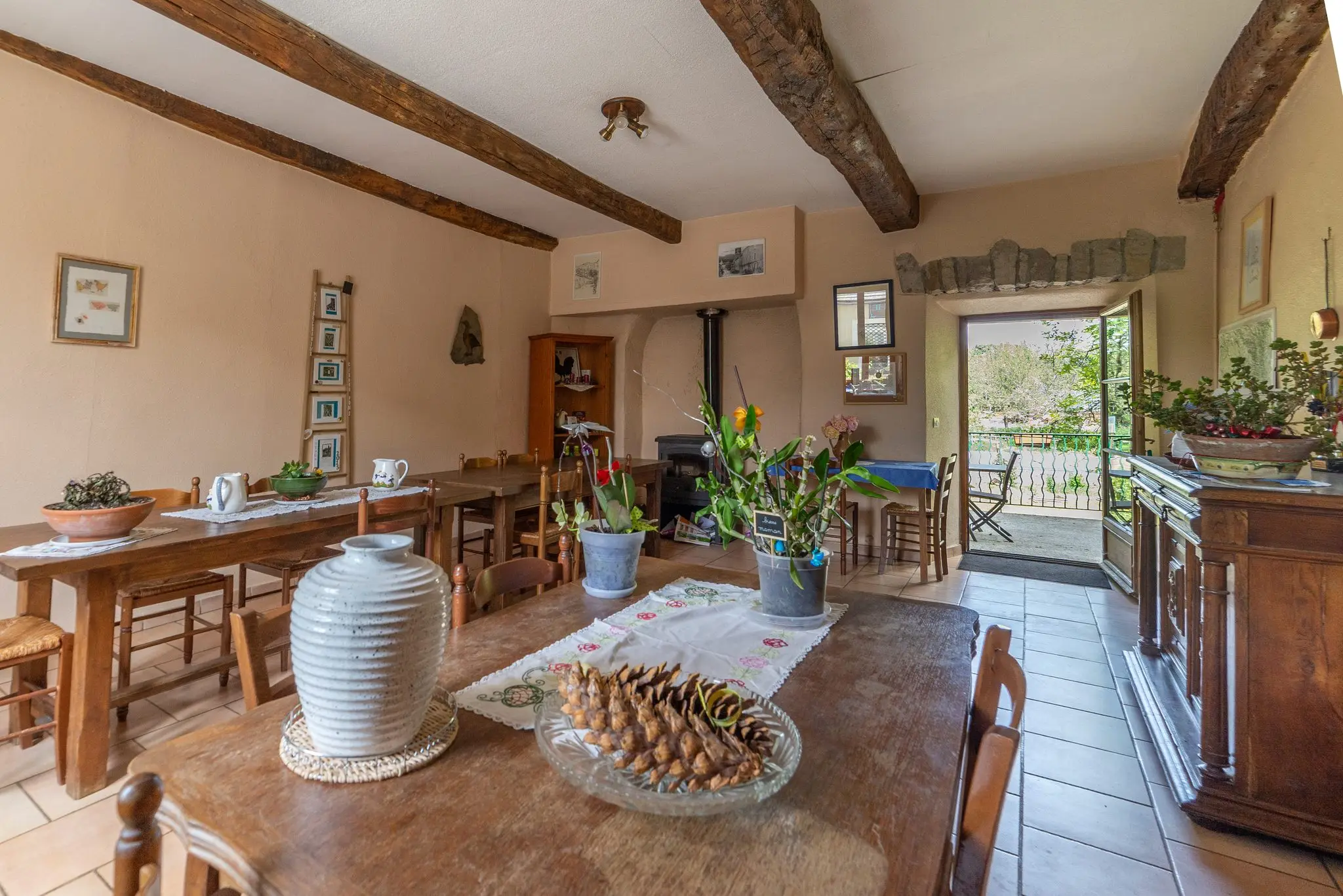 Grande salle commune aux poutres apparentes, longues tables en bois, sol en tomettes et porte vitrée ouverte sur la terrasse arborée