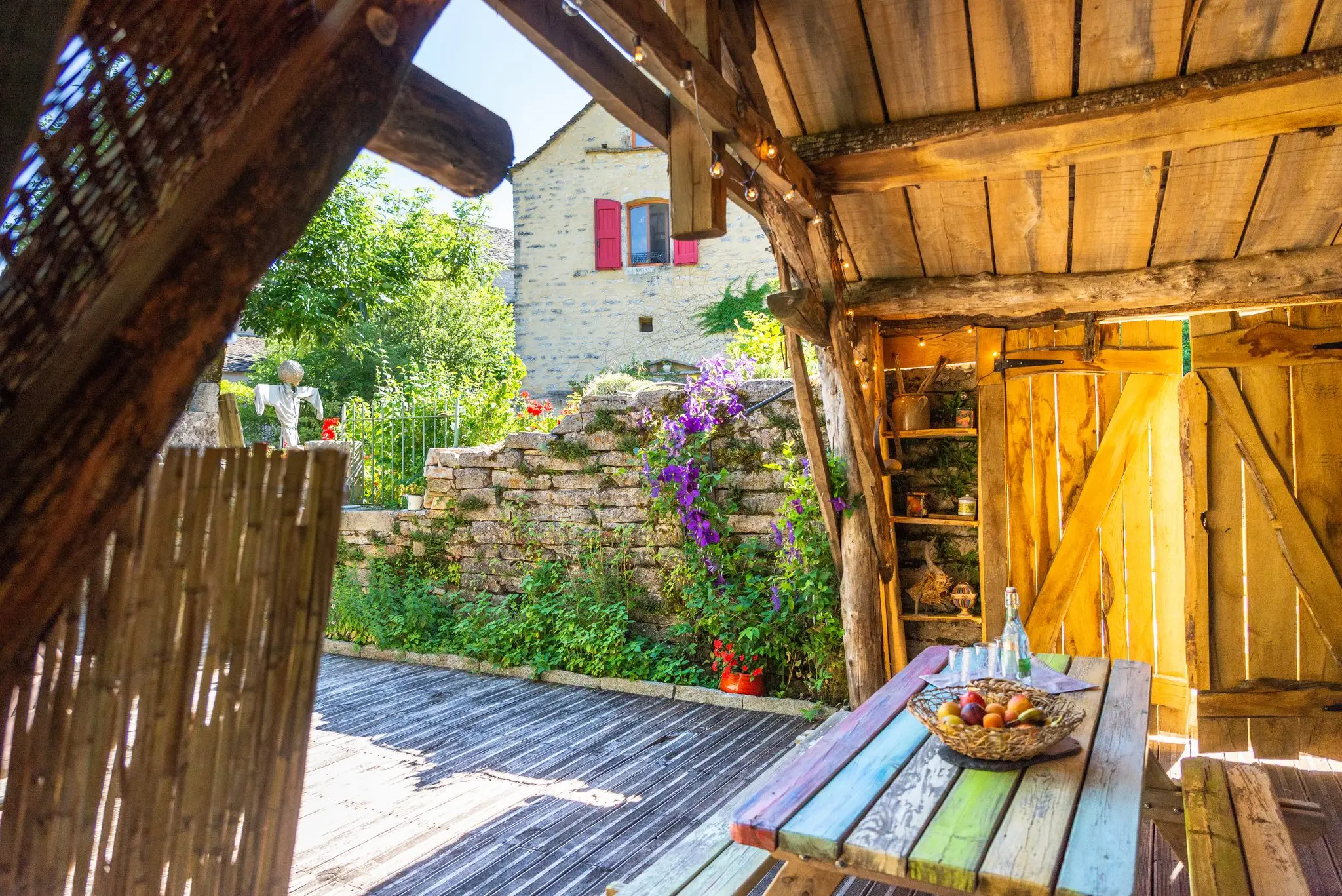 Terrasse couverte en bois d'un gîte, table colorée et vue sur une maison de pierre du hameau